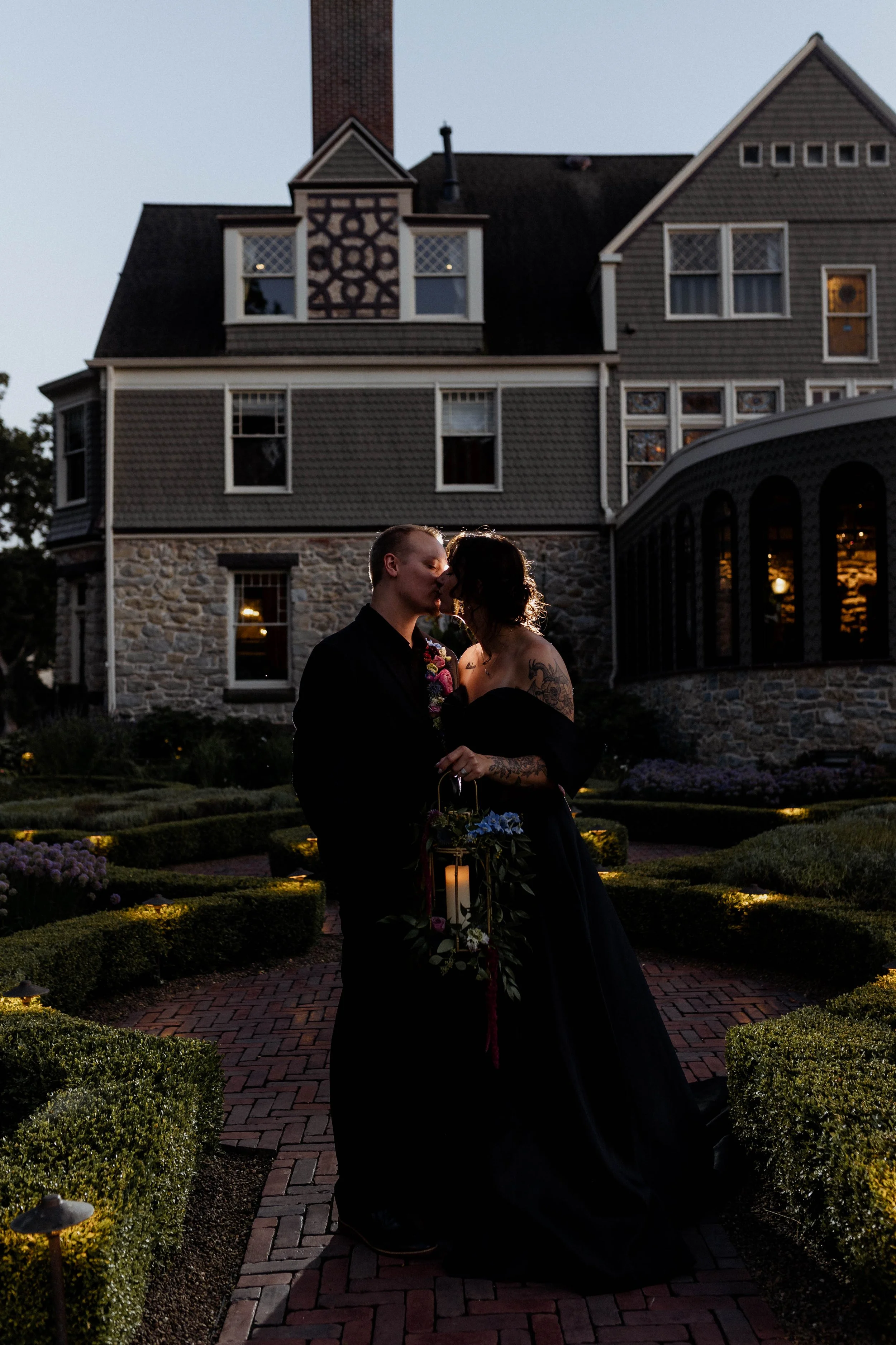 A couple kissing in front of a large house at dusk, with one holding a lantern decorated with flowers.