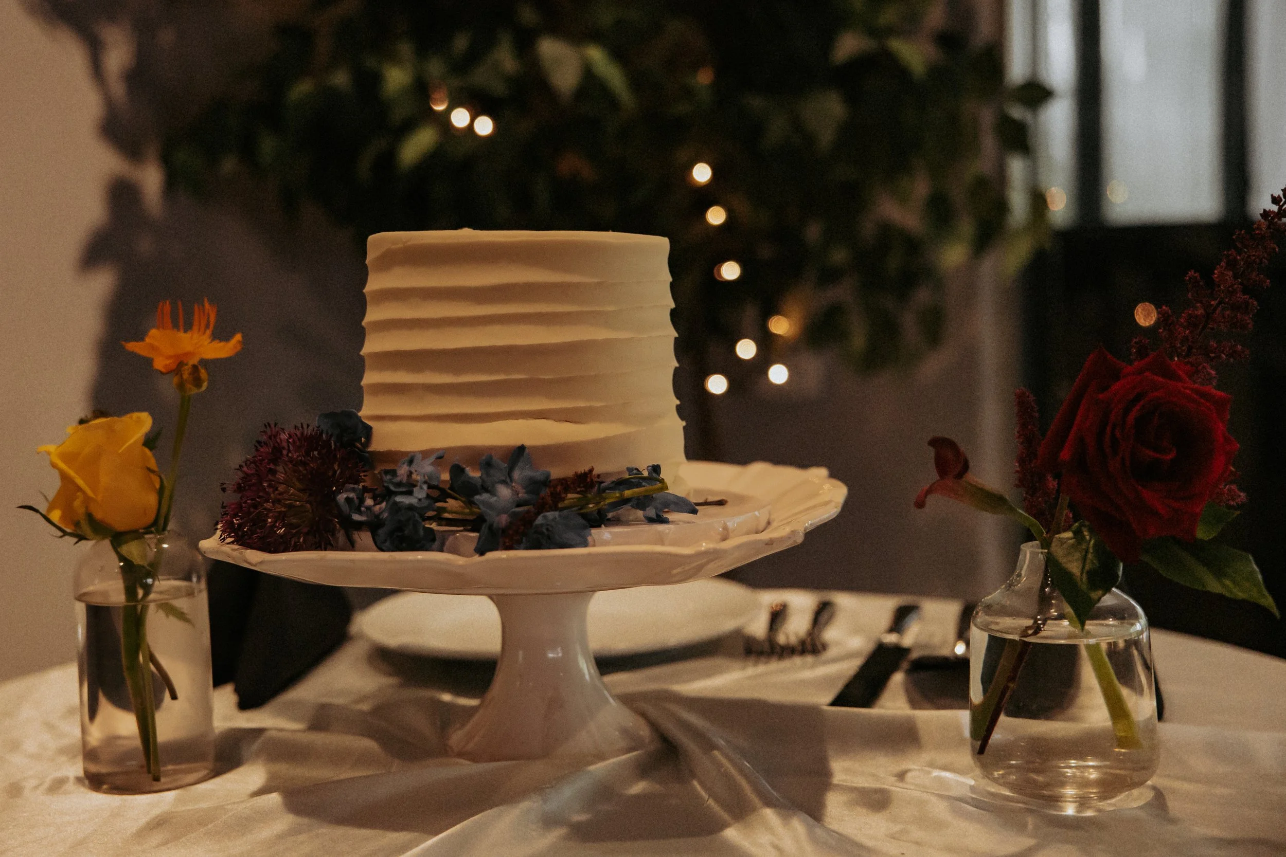 A white frosted cake with horizontal lines sits on a decorative white cake stand. The cake is adorned with dark purple and blue flowers. On each side of the cake, there are small glass vases with flowers, one with yellow roses and the other with a re