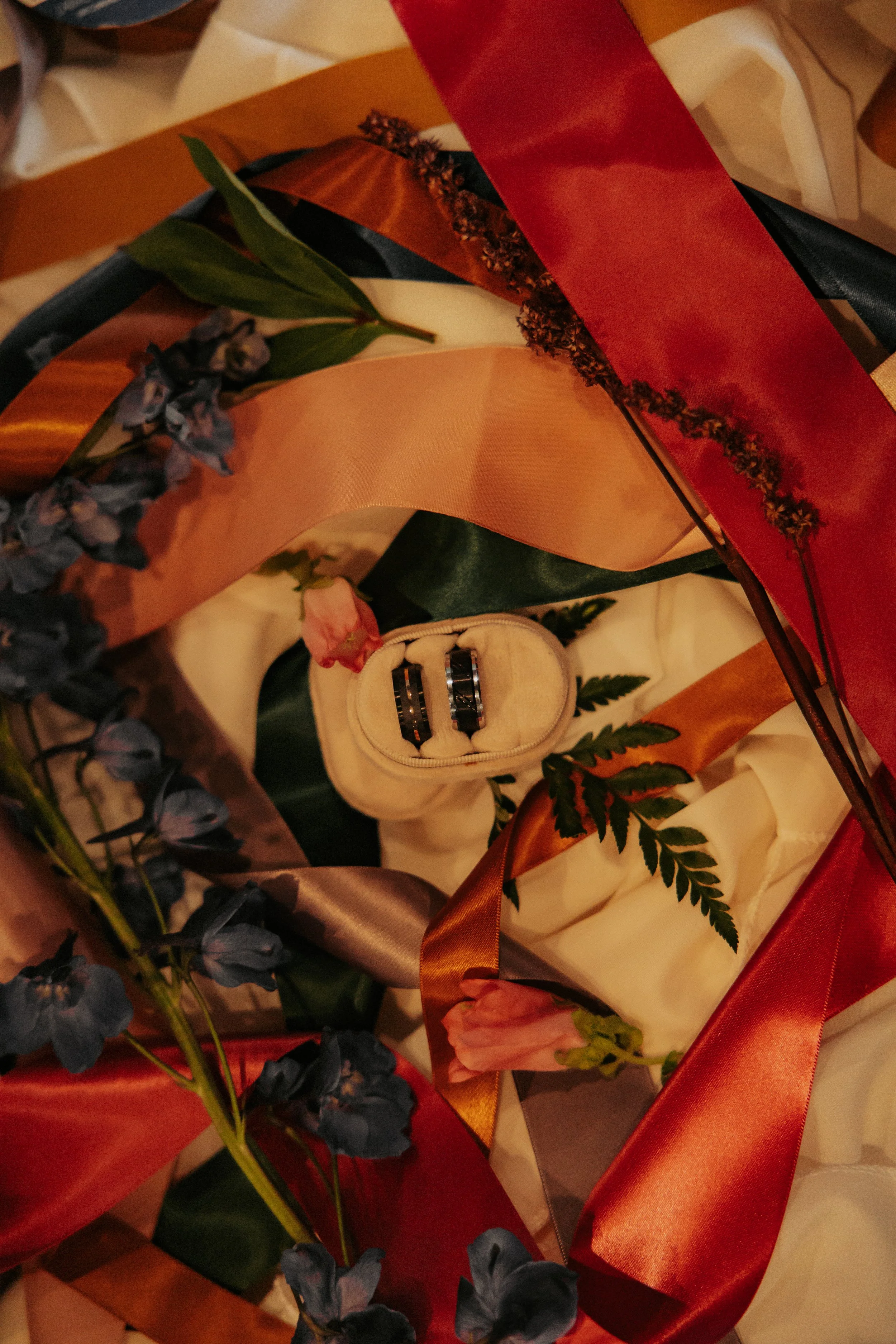 A gift box is decorated with colorful ribbons, flowers, and green leaves. Inside the box, there is a pair of rings in a small beige pouch.