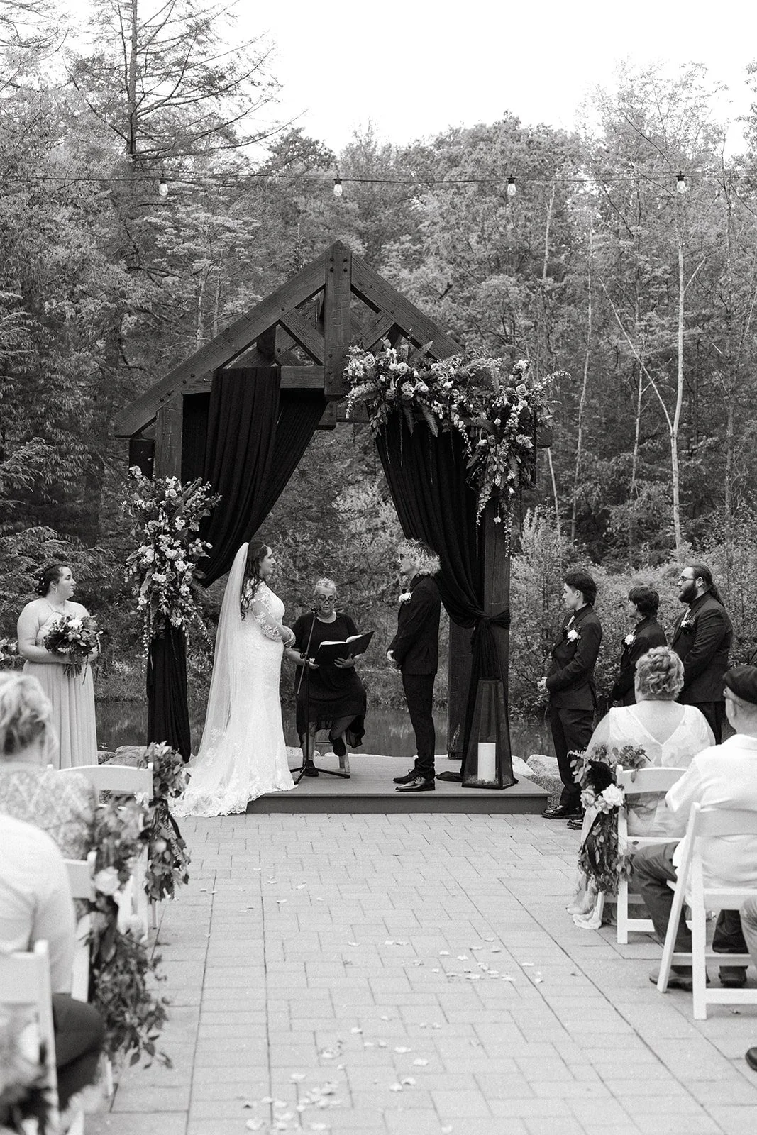 A black and white photo of an outdoor wedding ceremony with a wooden arch decorated with flowers. The bride and groom stand facing each other, with an officiant seated behind them, surrounded by guests seated on either side.