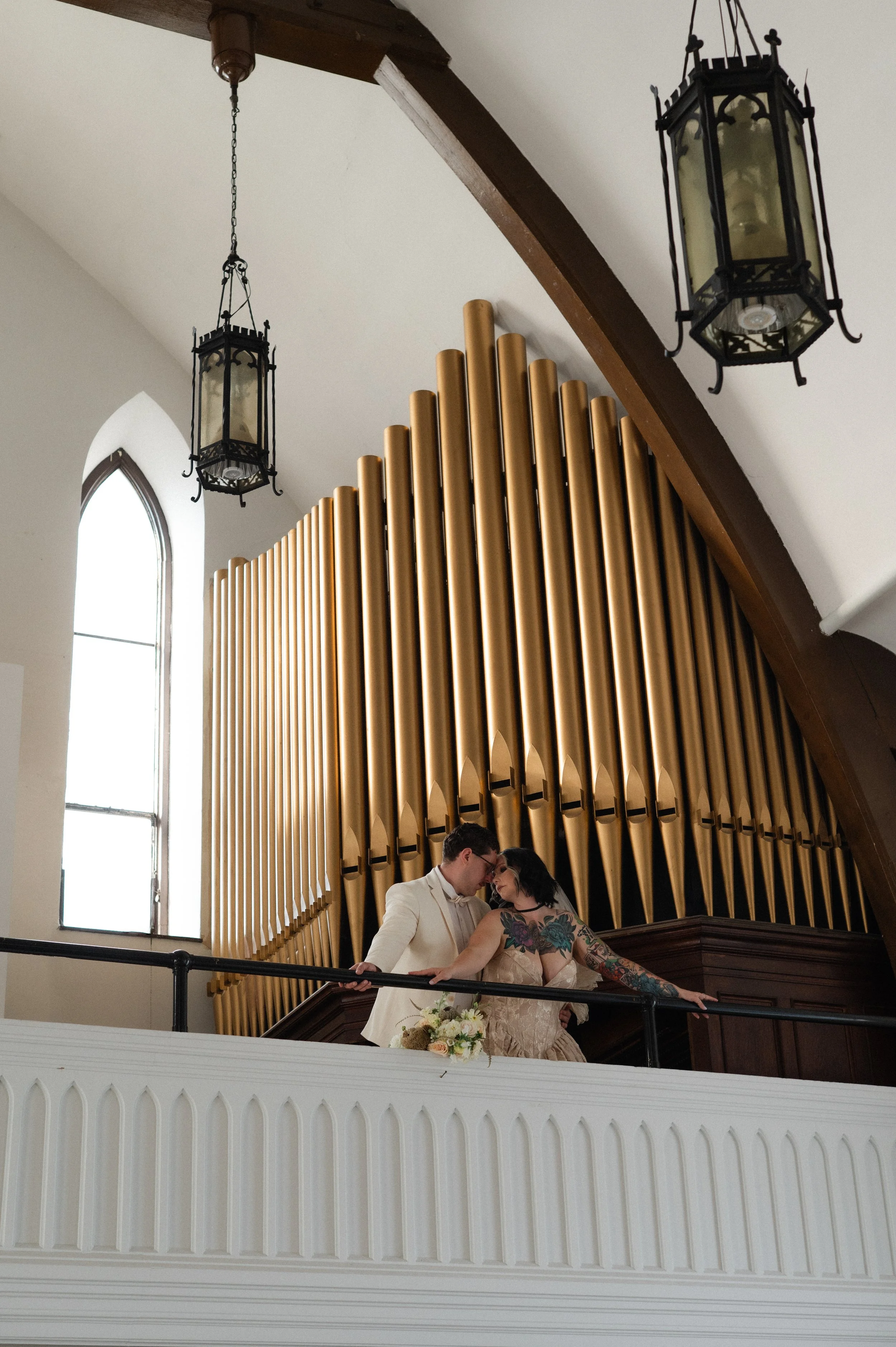 A couple embracing on a balcony with a large organ in the background, inside a church with tall arched windows and hanging lanterns.