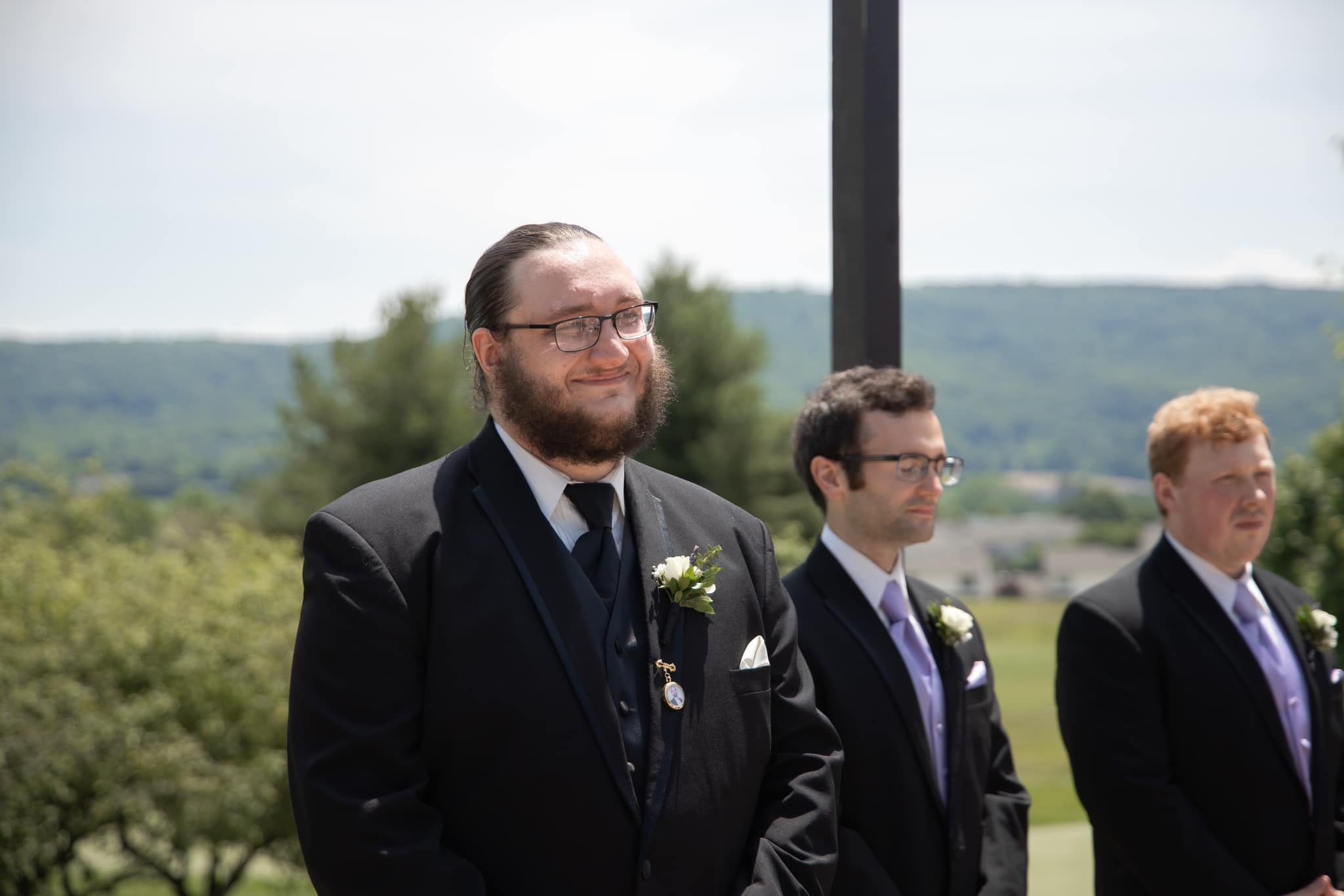Three men in formal suits attending an outdoor wedding ceremony. The man in the foreground has a beard, glasses, and is smiling gently. The other two men, standing behind him, wear glasses and have serious expressions. All three have white boutonnièr