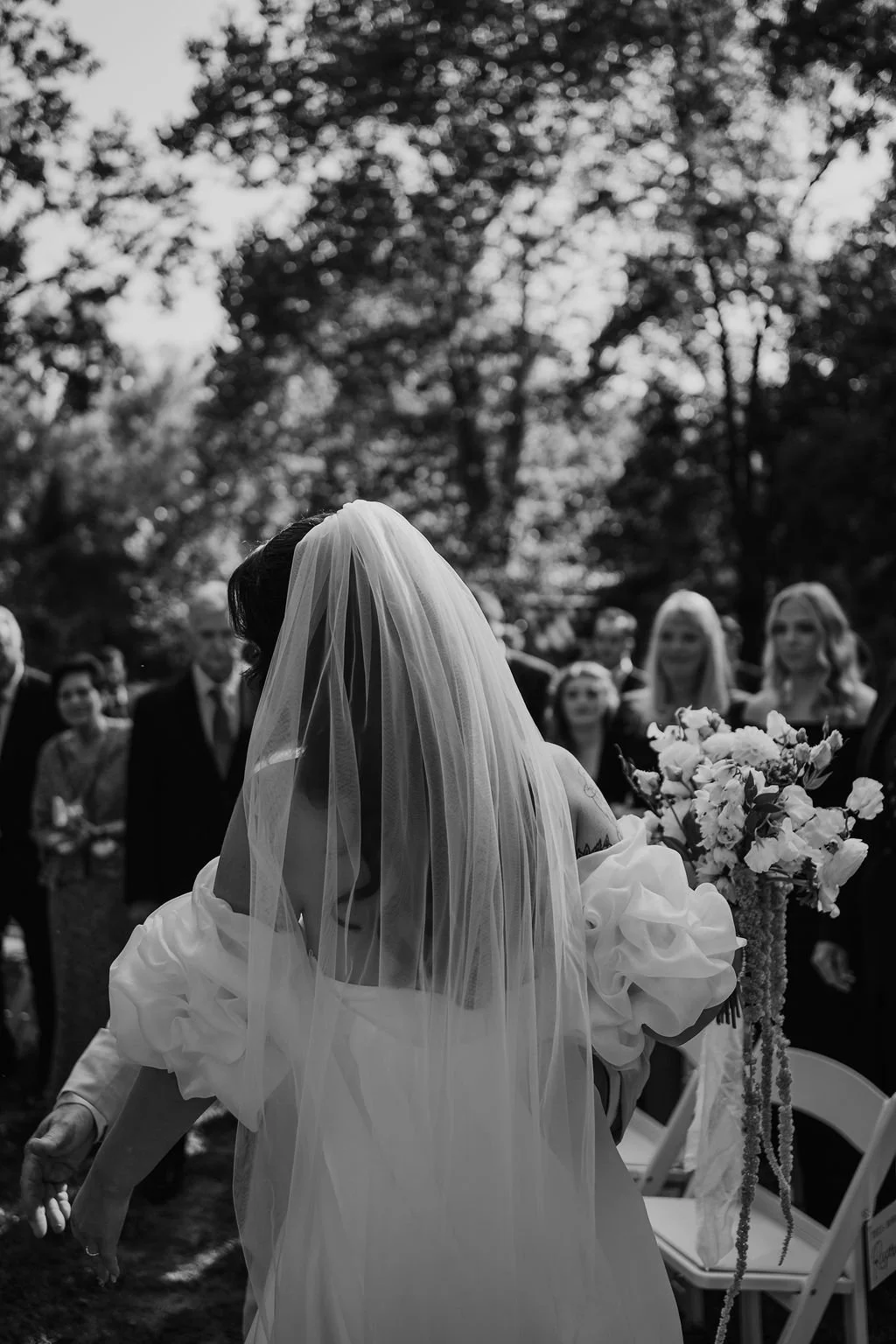 A bride with a veil and a flowing dress is holding a bouquet of flowers at an outdoor wedding ceremony. Guests are gathered in the background under trees.