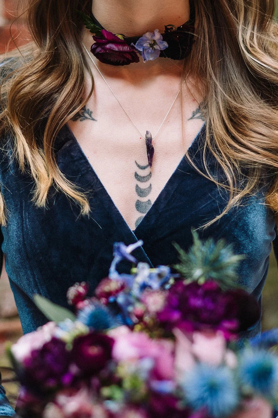 A woman wearing a crescent moon tattoo on her chest, a choker necklace with purple and black flowers, and a necklace with a purple crystal pendant, holding a colorful bouquet of flowers.