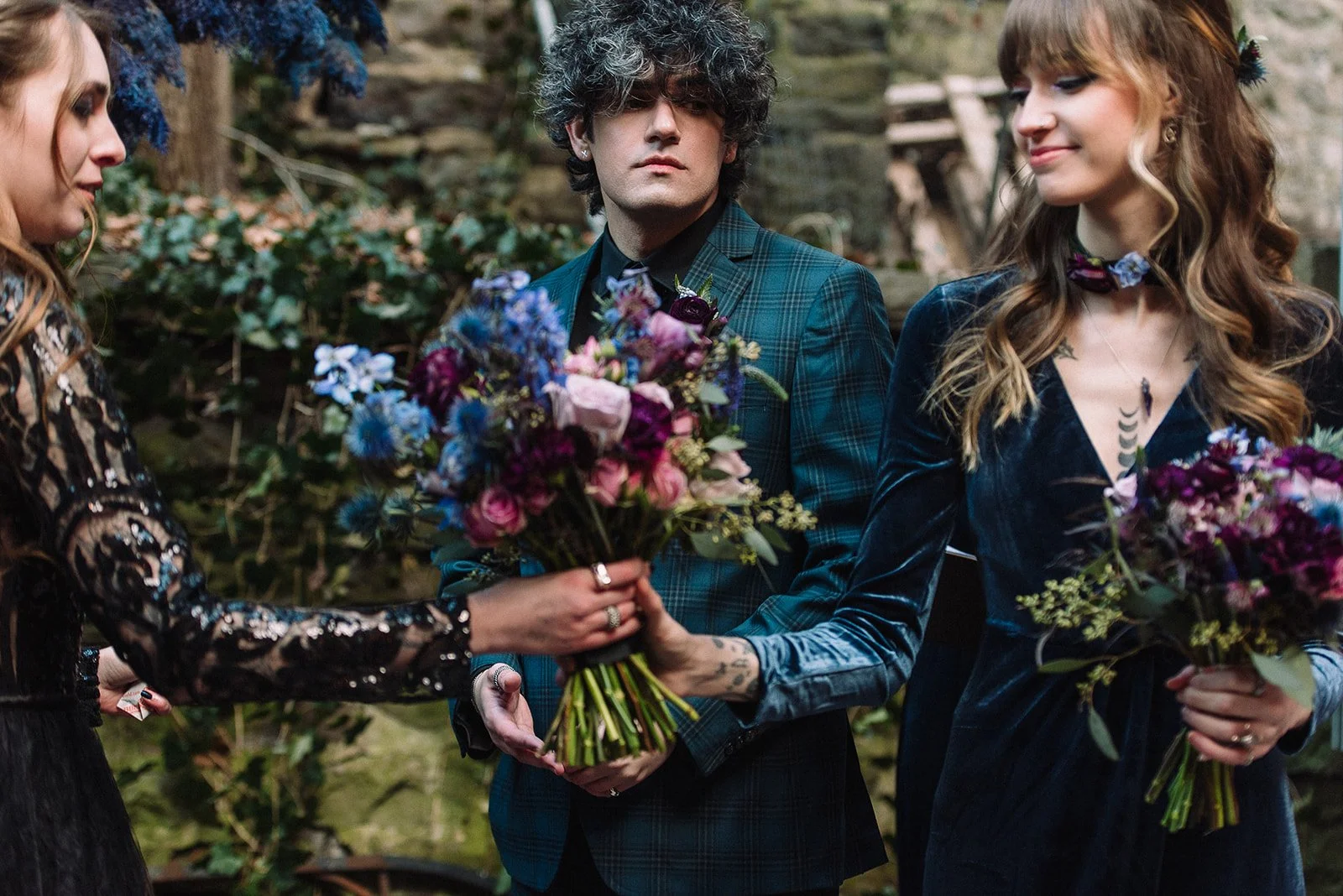 Three people, two women and one man, standing outdoors with a lush background of plants and trees. The women are holding colorful bouquets of flowers and smiling, while the man is looking at the camera with a neutral expression.