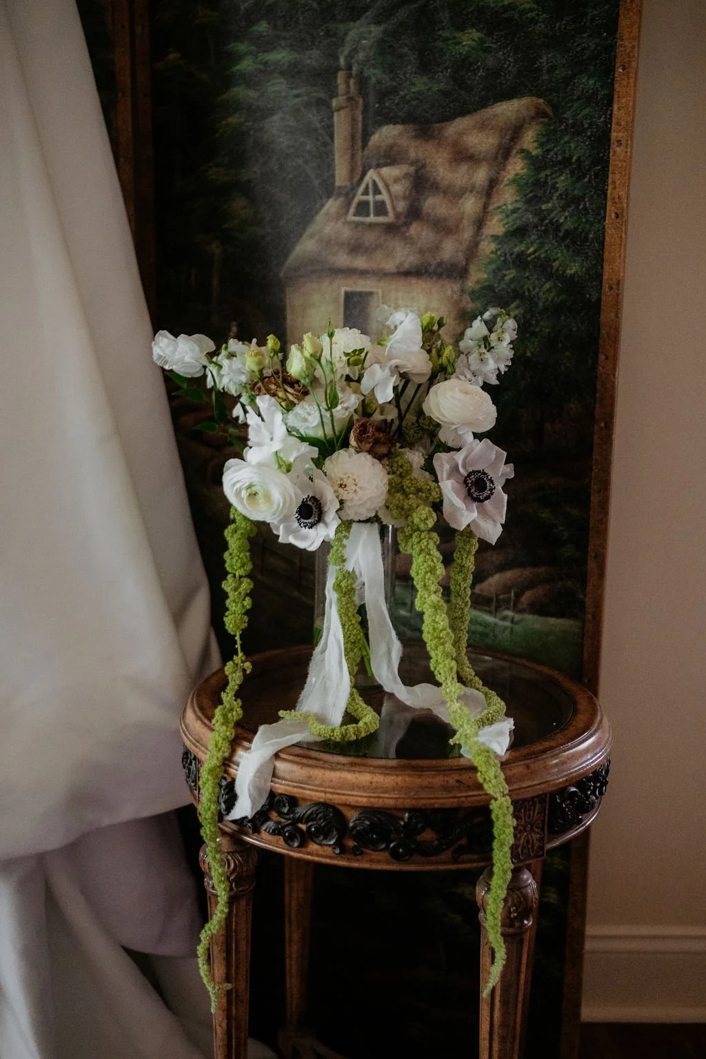 A bouquet of white flowers with green hanging accents in a glass vase, placed on an ornate wooden side table with a painting of a cottage and forest in the background.