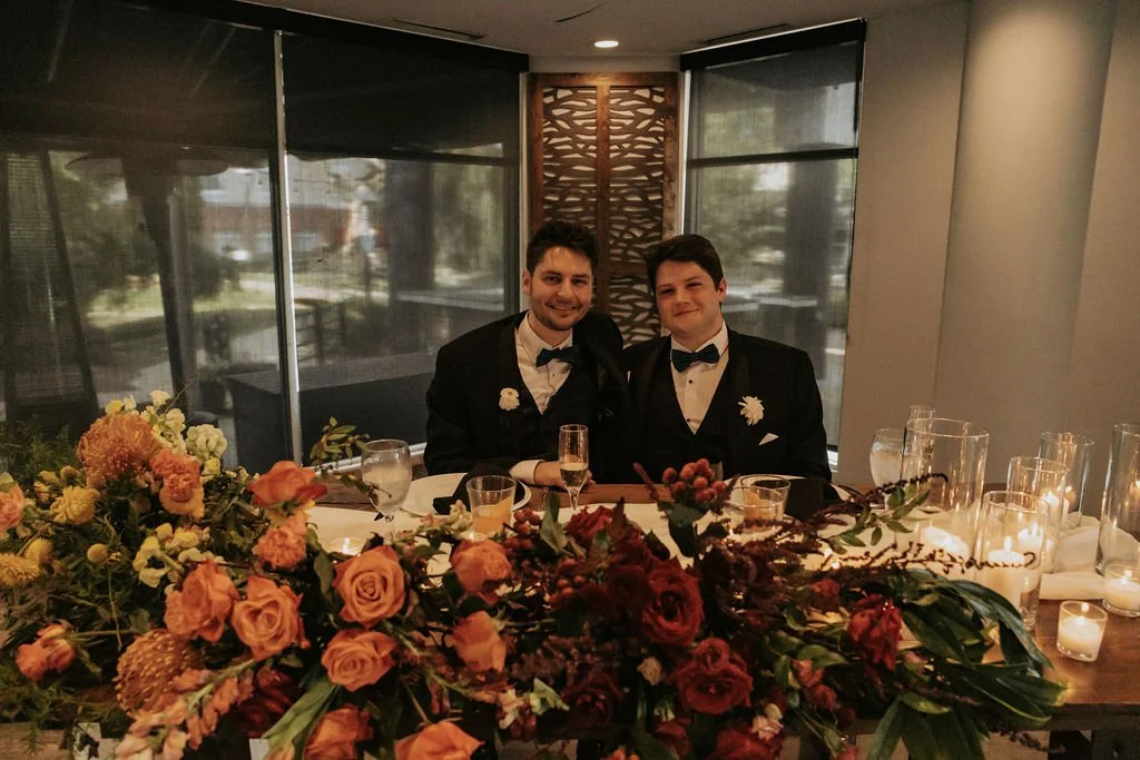 Two men dressed in tuxedos and bow ties sitting at a decorated table with floral arrangements and candles, smiling for the camera in a dimly lit, elegant setting.