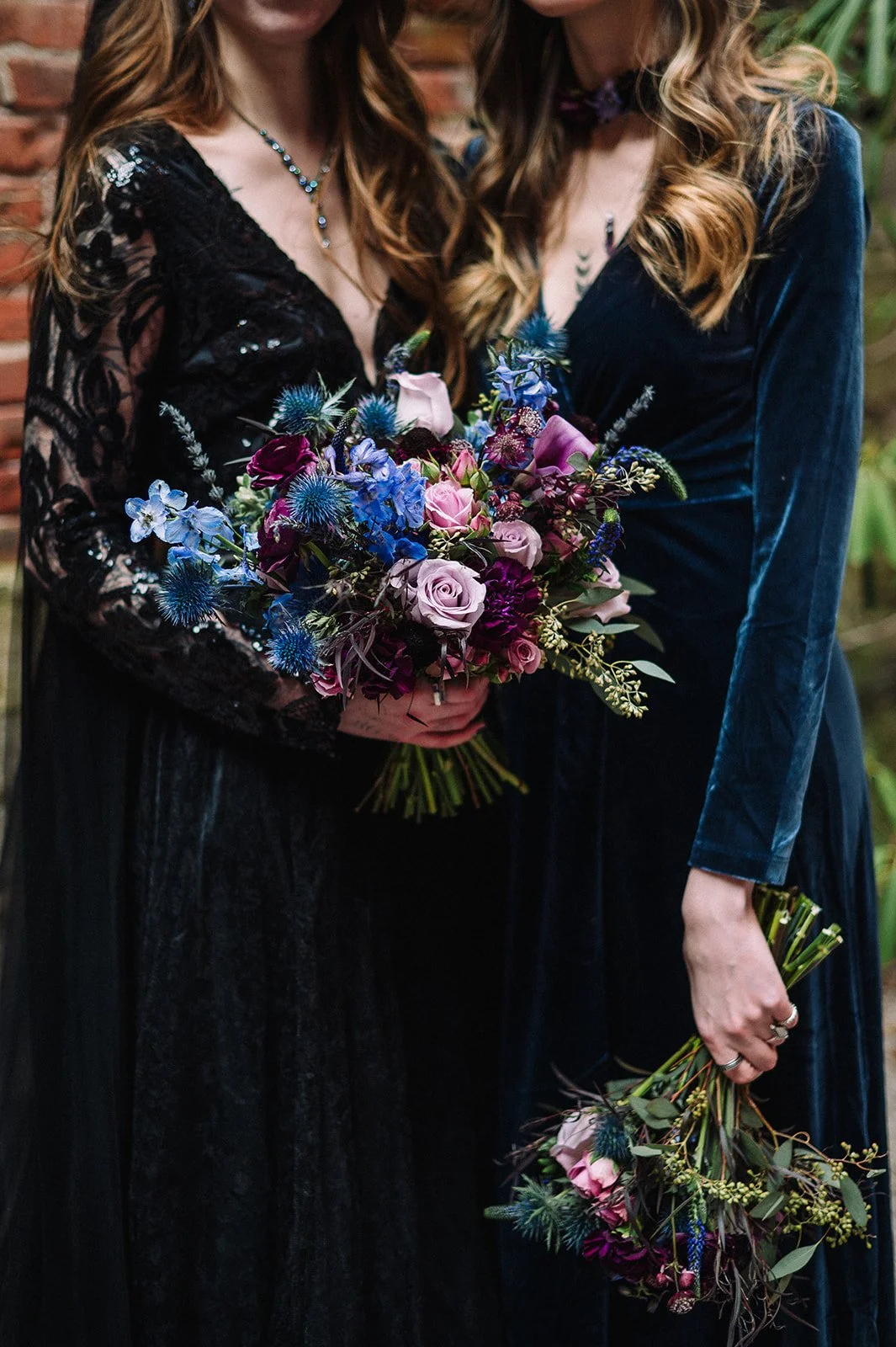 Two women wearing dark dresses holding bouquets of purple, pink, and blue flowers, standing close together outdoors.