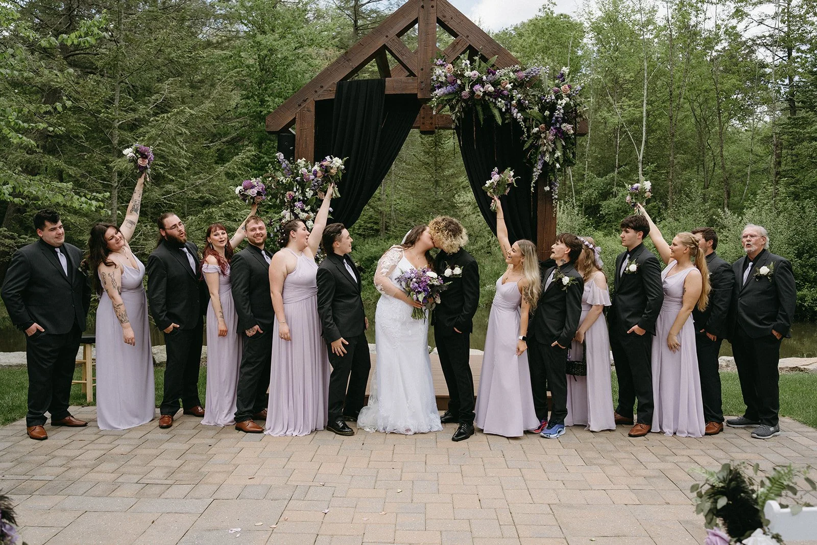 A wedding ceremony outdoors with a bride and groom kissing, surrounded by their wedding party, under a wooden arch decorated with flowers and black drapery, in a lush green setting.