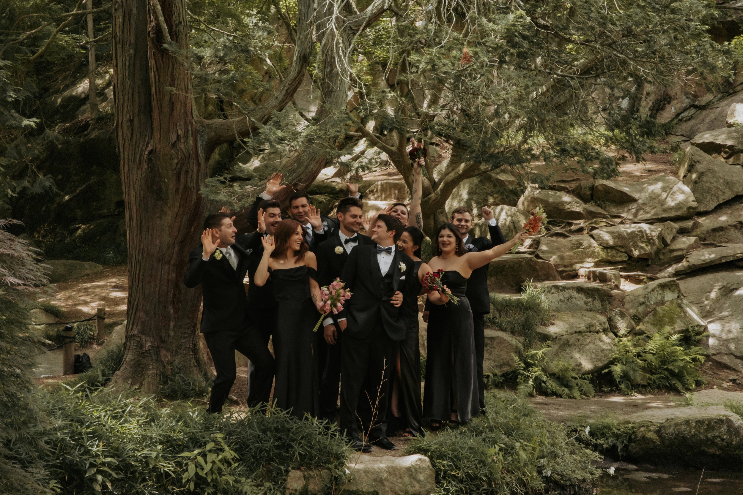 A group of people in formal attire celebrating outdoors under a large tree and rocks.