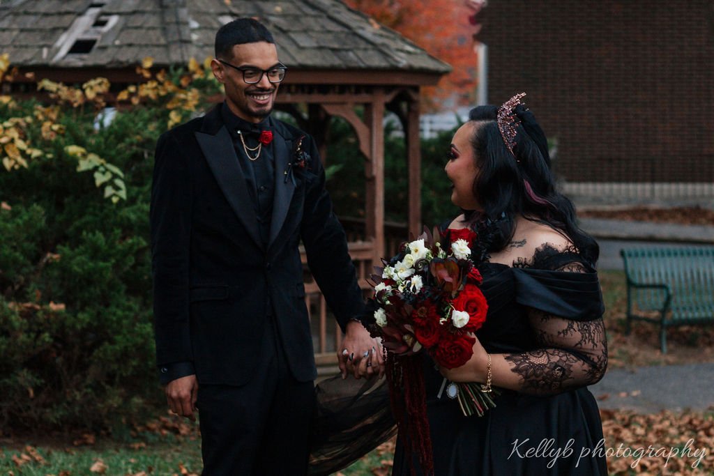 A man and woman dressed in formal attire holding hands outdoors, with the woman holding a bouquet of red and white flowers. The woman is smiling, and the man has a happy expression. The scene appears to be during fall, with colorful leaves and outdoo