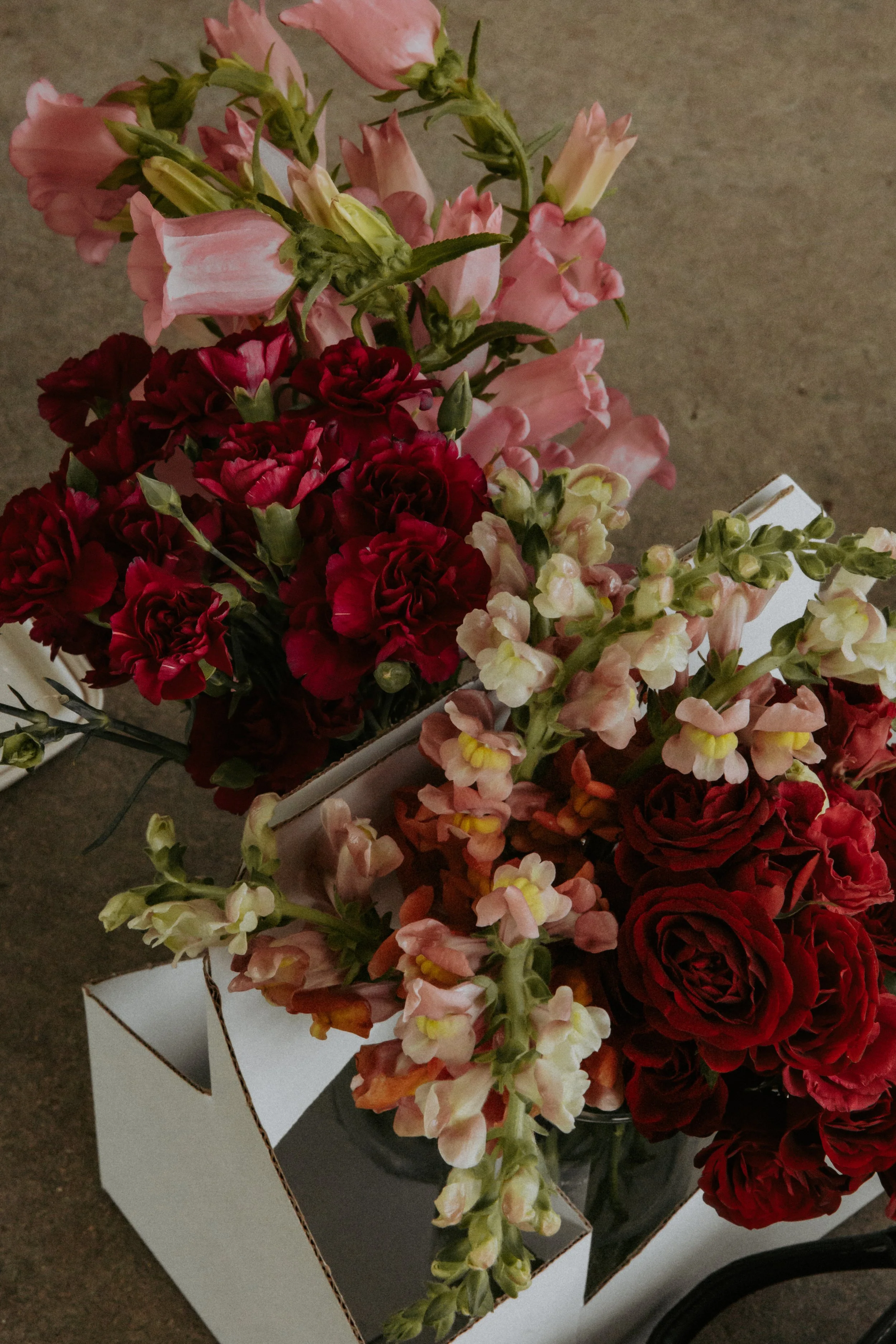 A bouquet of pink, red, and white flowers in a cardboard box on a concrete floor.
