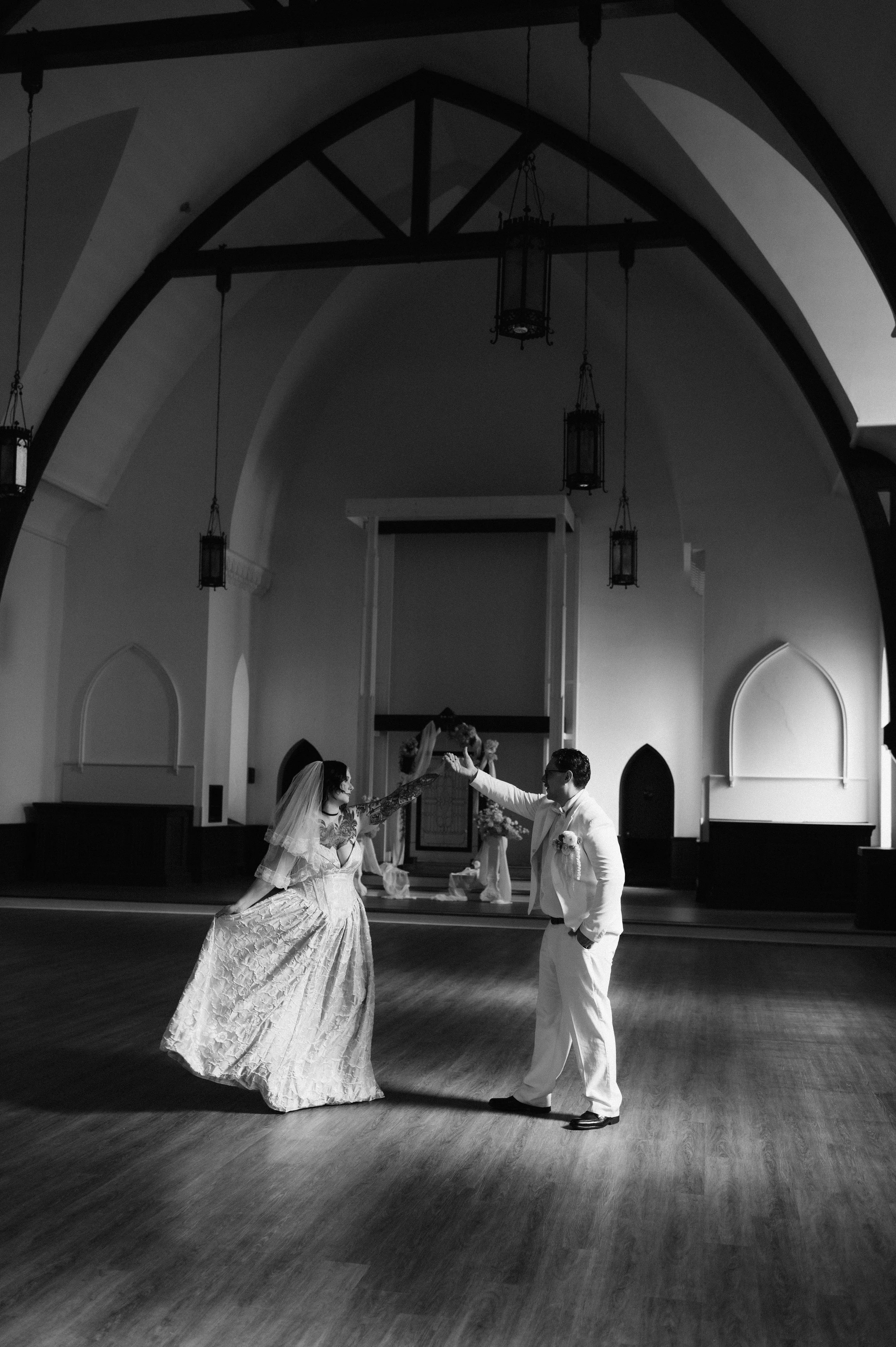 A black and white photo of a bride and groom dancing in a church, with the bride wearing a long gown and veil, and the groom in a white suit, under high arched ceilings with hanging lanterns.