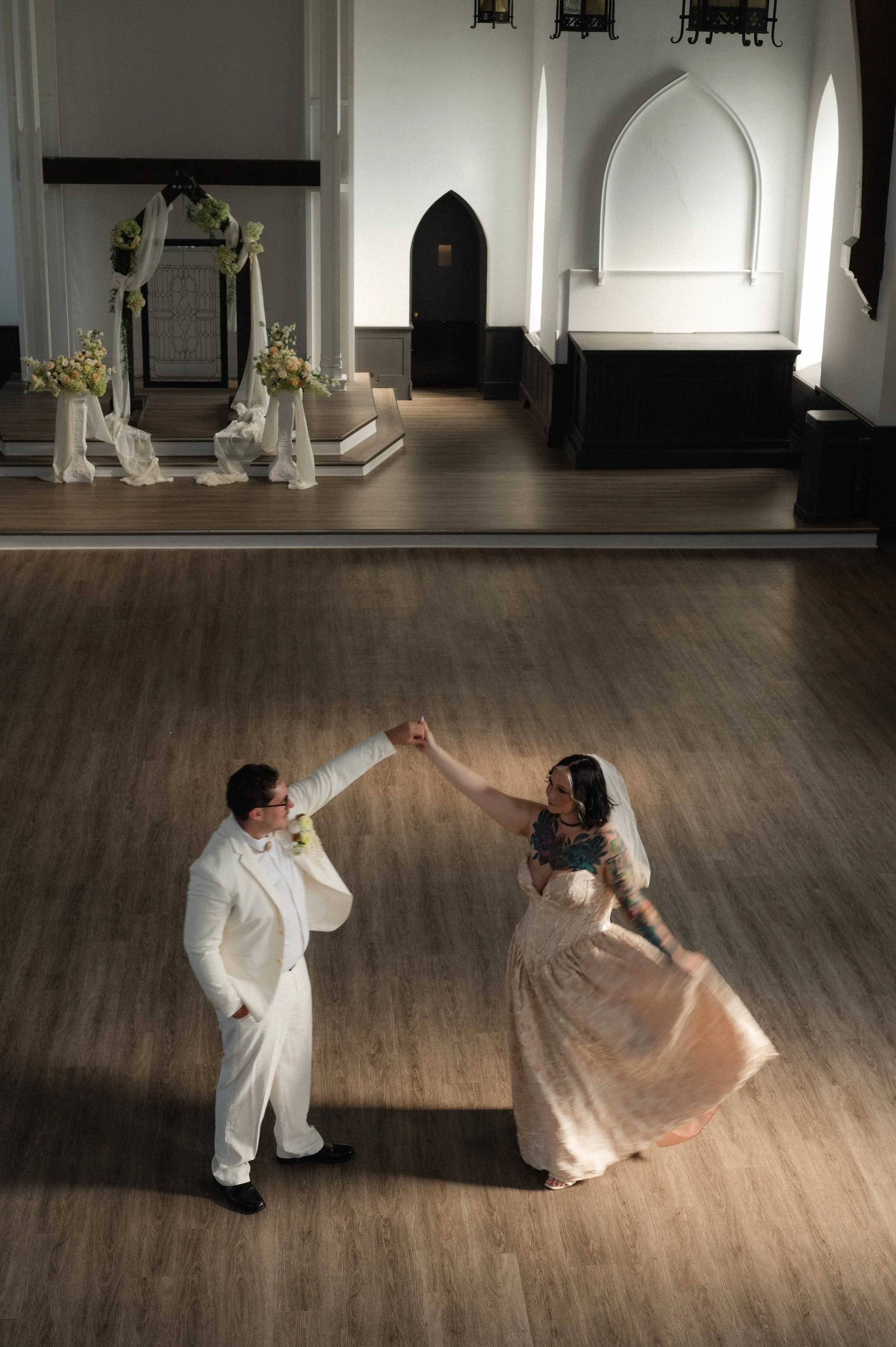 A bride and groom dancing in an empty church or wedding venue, with the bride spinning and the groom holding her hand, in front of an altar decorated with flowers and fabric.