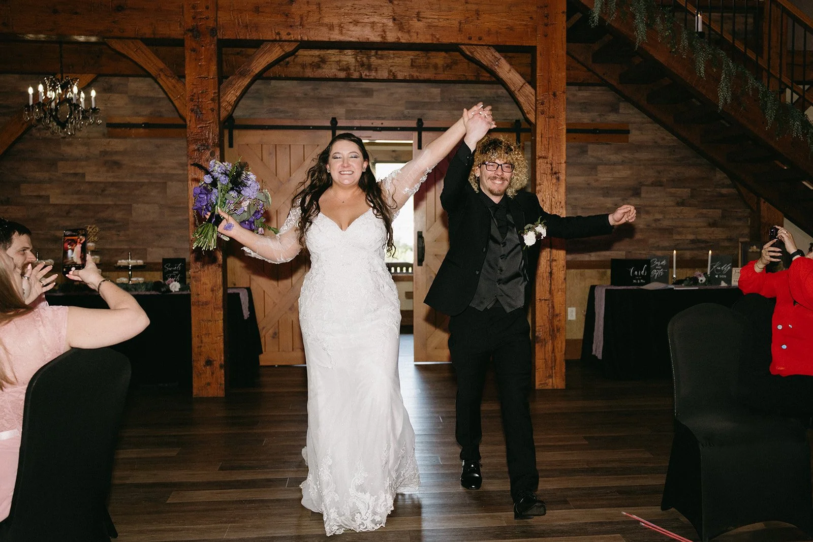 A newlywed couple walking into a reception venue, holding hands and smiling. The bride is in a white lace wedding gown holding a purple bouquet, and the groom is in a black suit with glasses. Guests are taking photos on either side.