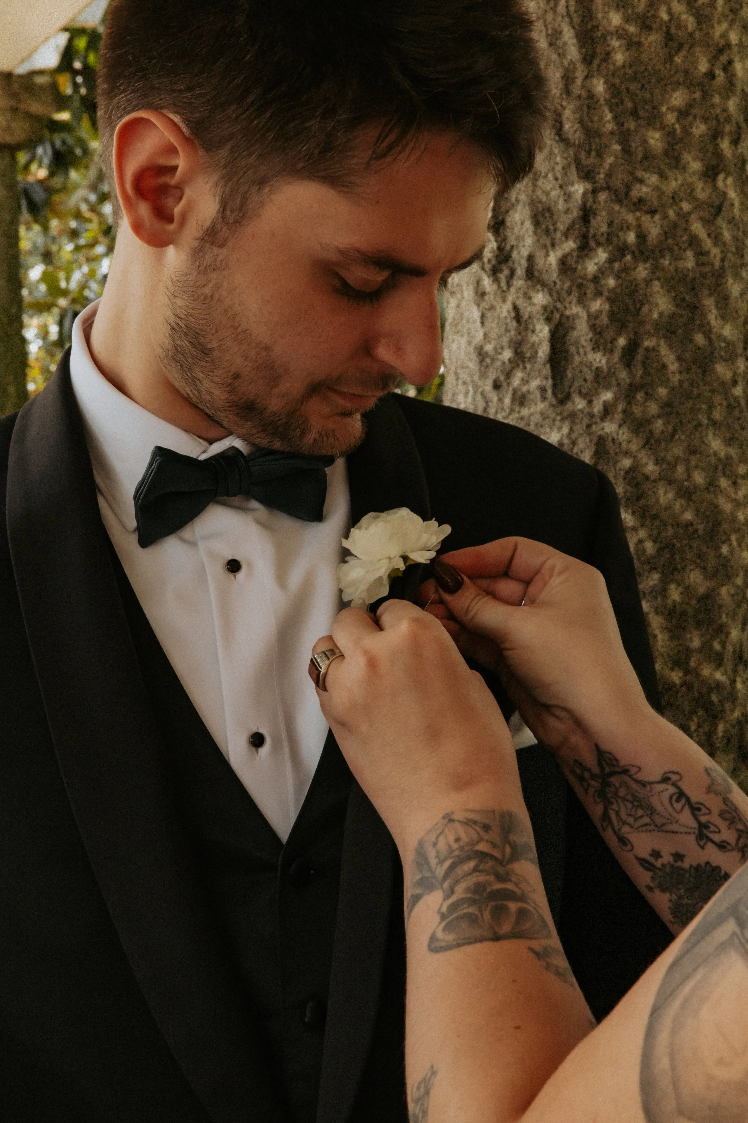 A groom in a black tuxedo with a bow tie is being assisted in pinning a white flower boutonniere on his lapel by a person with tattoos on their arms.