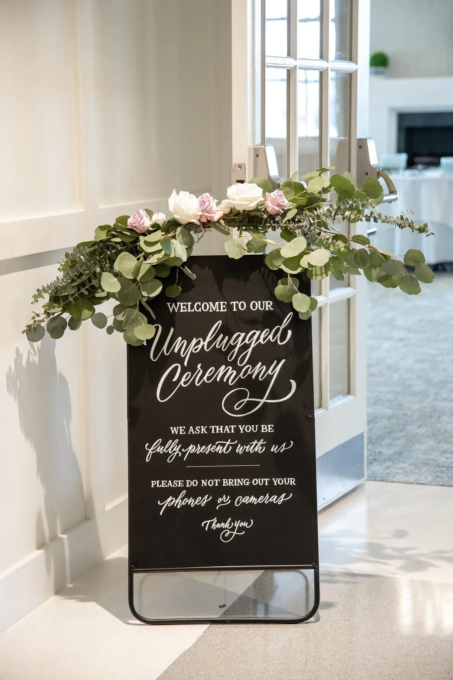 A black welcome sign with white cursive and print text reading "Welcome to our Unplugged Ceremony," decorated with pink and white flowers and eucalyptus leaves, placed at the entrance of an event space.