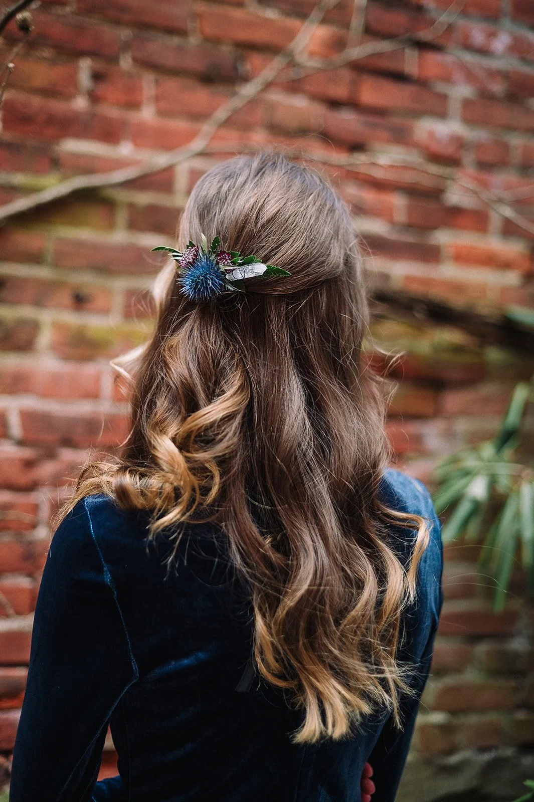 Back view of a woman with long, curly brown hair, decorated with a blue thistle flower and greenery clip, wearing a dark blue velvet jacket, standing outdoors near a brick wall.