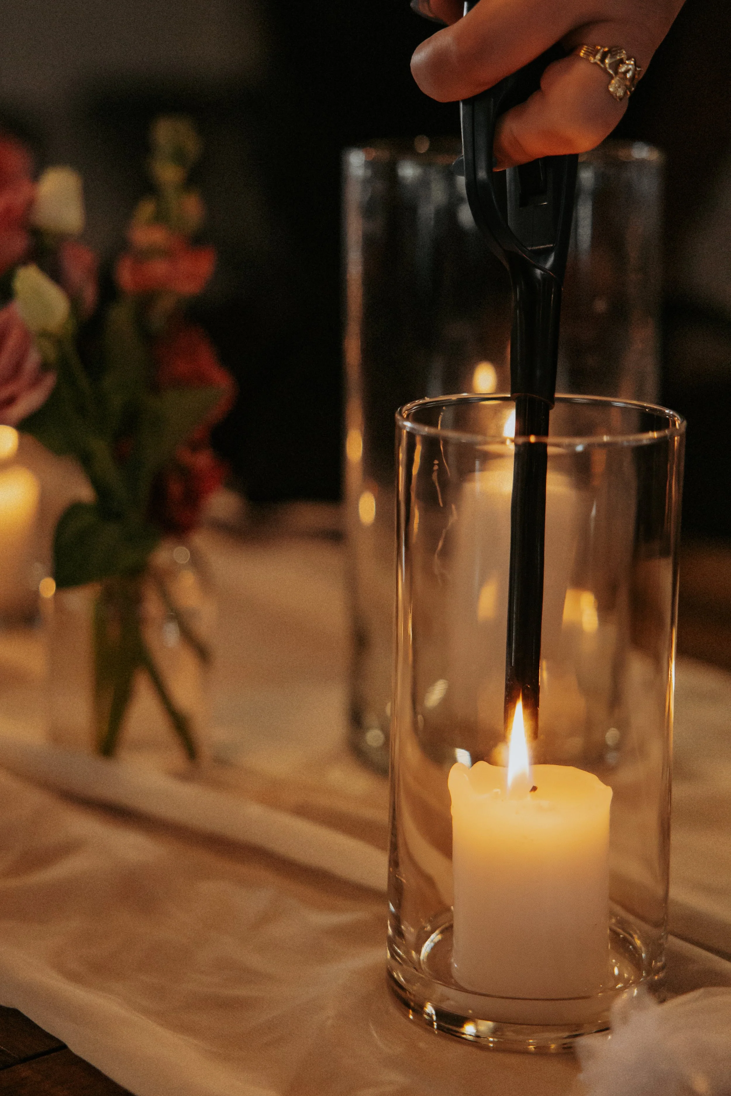 A hand placing a black table lighter near a lit white candle inside a glass holder, on a table with a white table runner and blurred decorative items in the background.