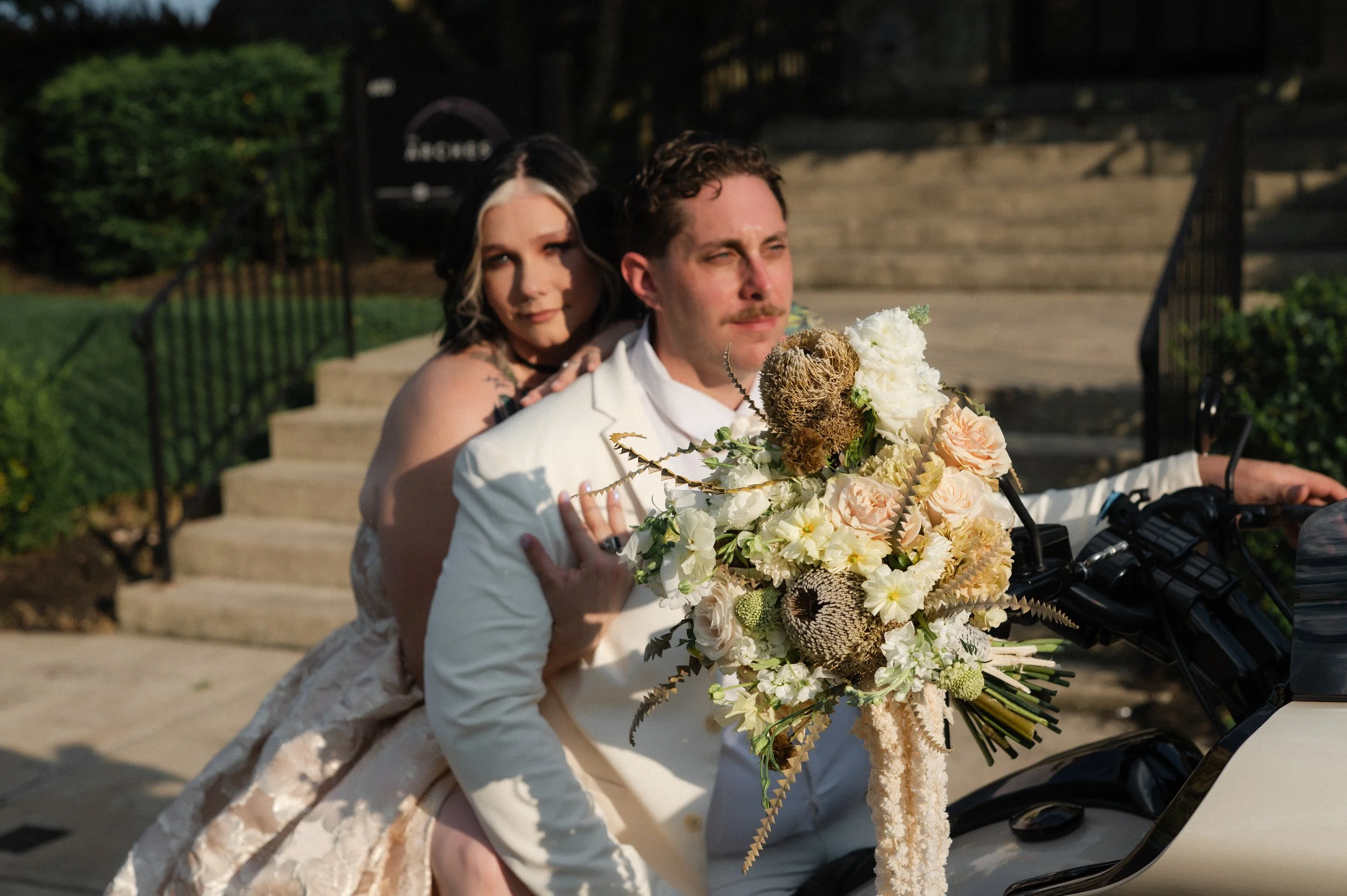 A couple, dressed in wedding attire, on a vintage motorcycle with a large bouquet of flowers in front of them. The man is wearing a white suit with a boutonnière, and the woman is in a lace dress with dark hair and blonde highlights, both looking int