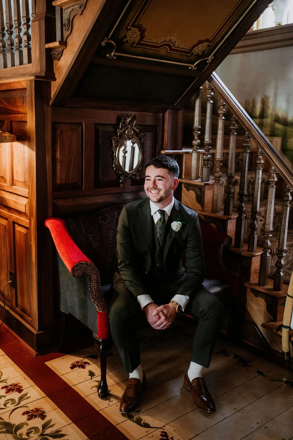 A smiling man in a suit sitting on a vintage red and black armchair in a wooden room with a staircase, a mirror, and decorative wall art.