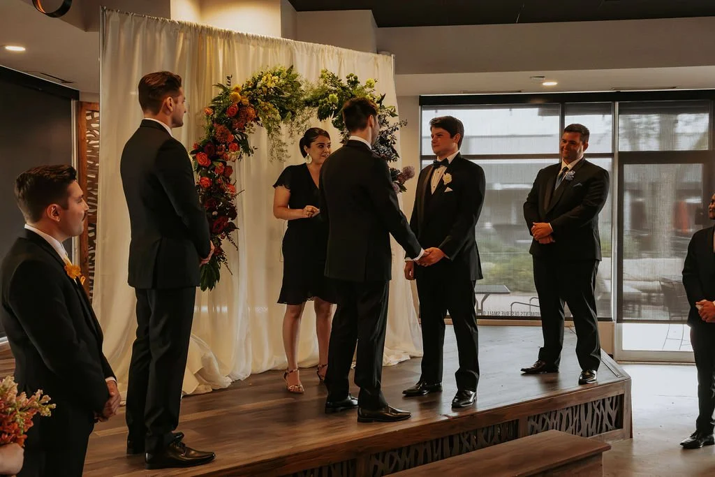 A wedding ceremony with a couple exchanging vows on a small stage, surrounded by groomsmen and a bridesmaid, inside a room with large windows and a floral backdrop.