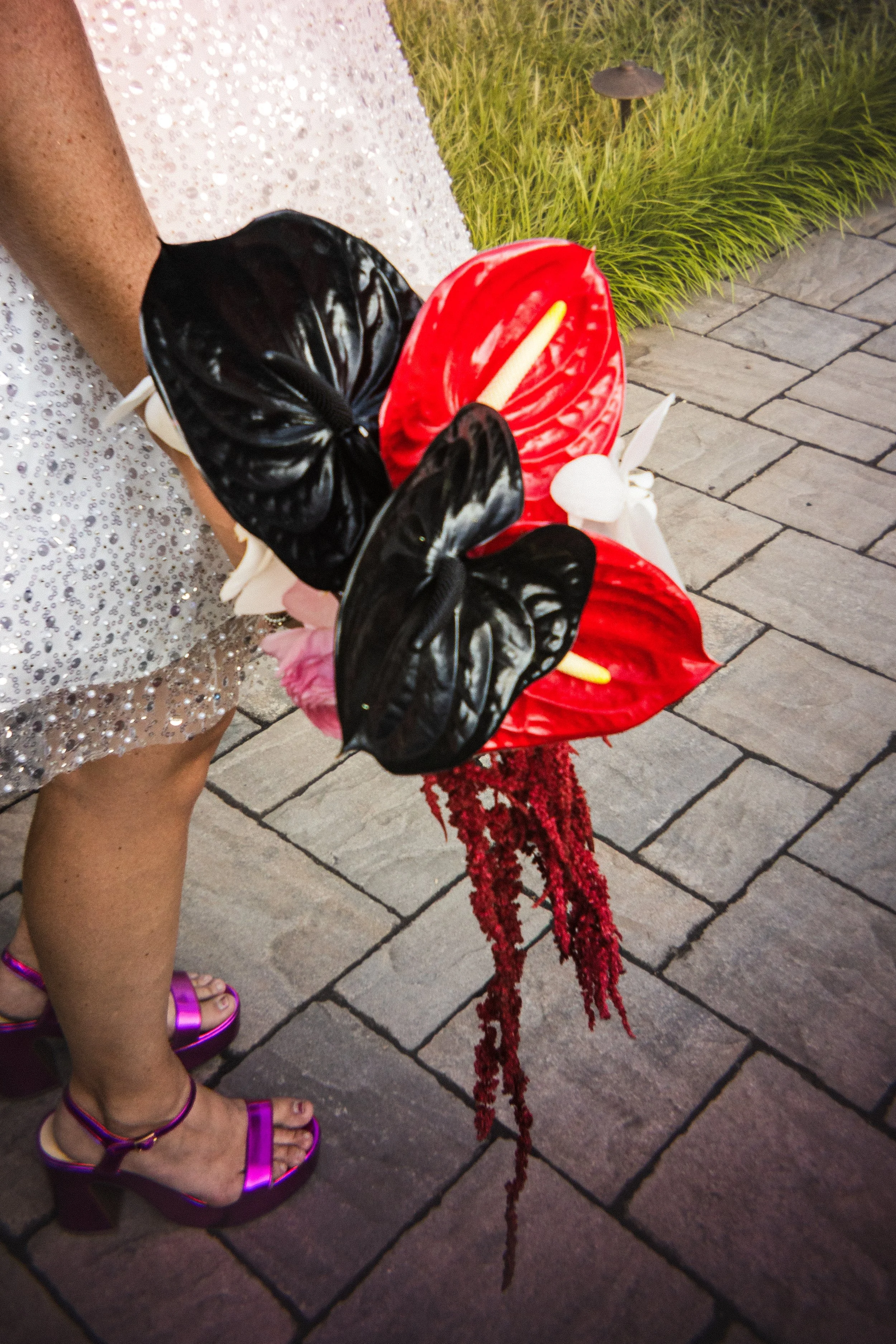 Person holding a bouquet of red and black anthurium flowers with pink and white accents, standing on a paved outdoor area.
