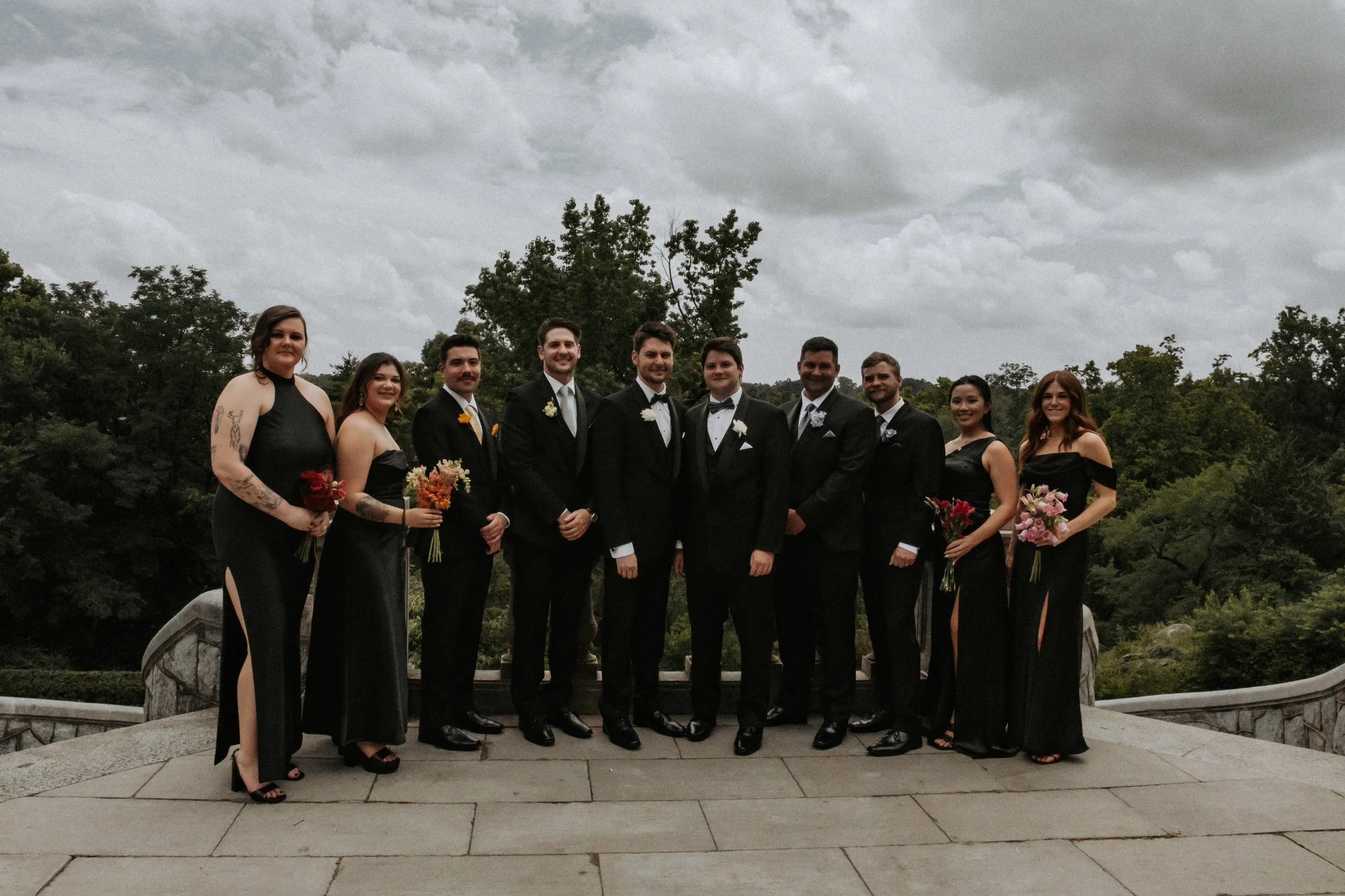 Group of wedding party members in formal attire standing outdoors on a stone terrace with a cloudy sky and trees in the background.
