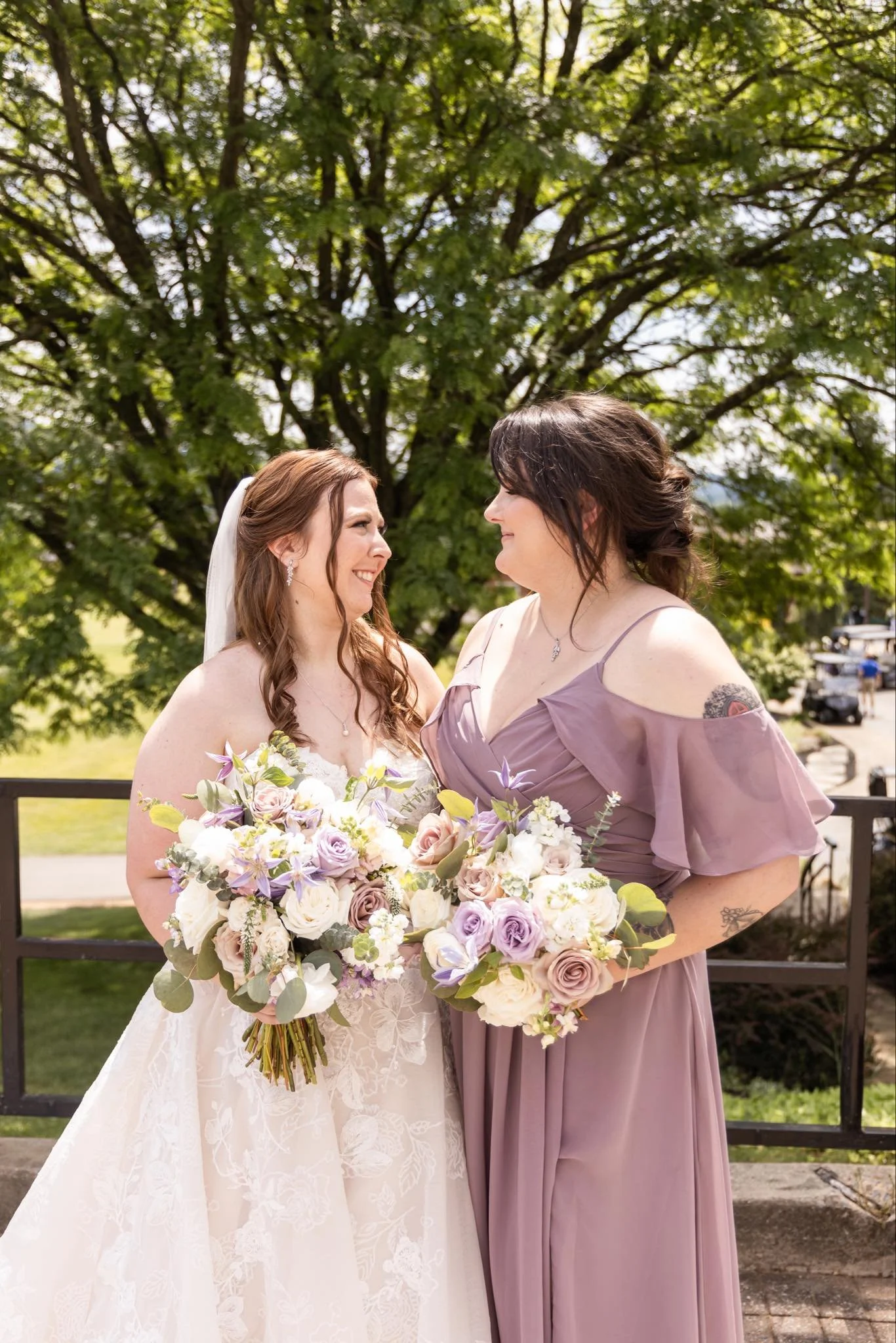 Two women, one in a white wedding dress and the other in a mauve bridesmaid dress, holding bouquets of flowers, standing close and looking at each other, outside with green trees in the background.