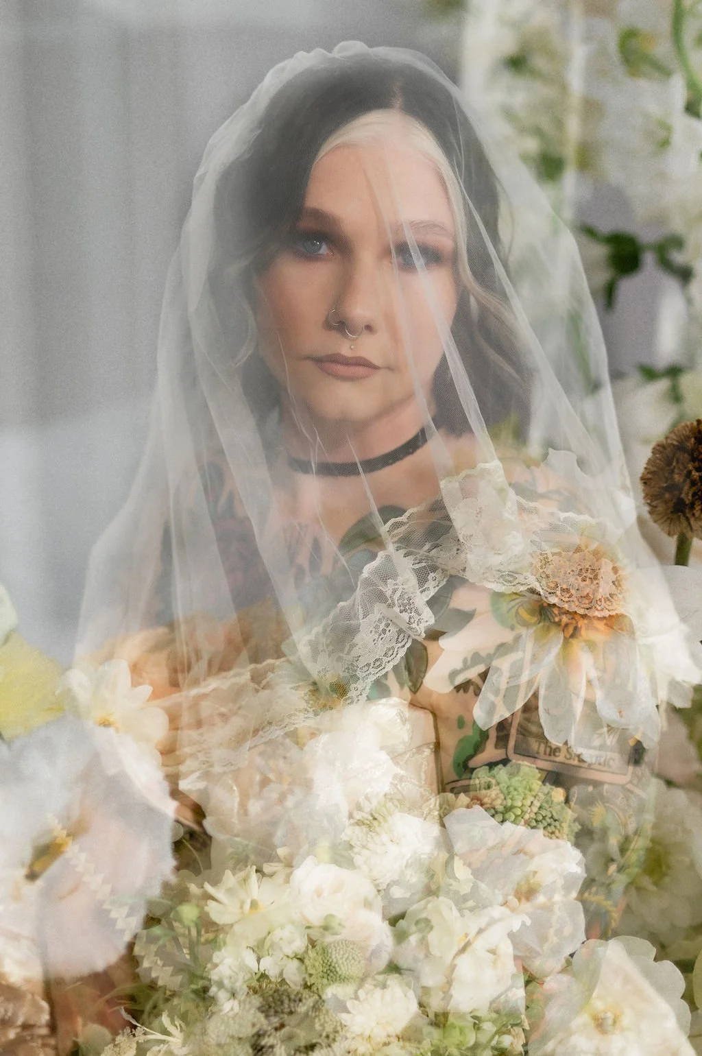 A woman with black and blonde hair, wearing a veil, choker, and colorful clothes, is surrounded by flowers and greenery.