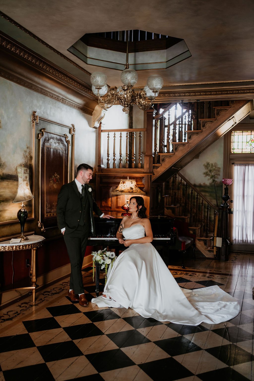 A bride and groom in vintage-style interior with staircase, chandelier, piano, and stained glass window, during wedding photoshoot.