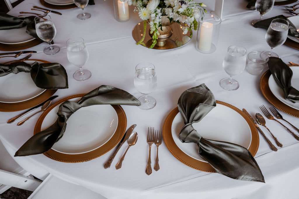 Elegant round dining table set for a formal event with white tablecloth, black napkins tied around white plates, gold chargers, drinking glasses, silverware, and a floral centerpiece with candles.