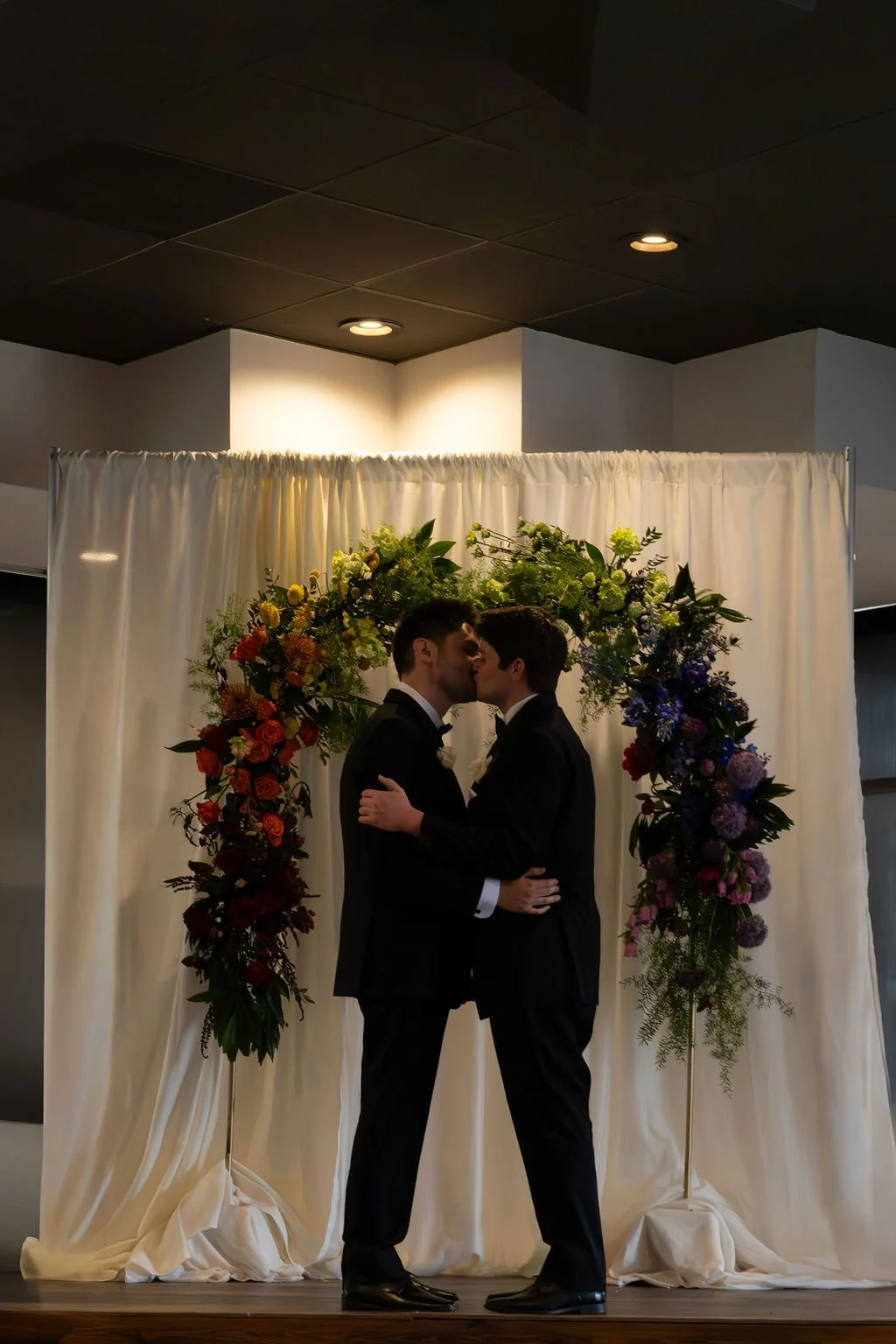 Two men in tuxedos sharing a kiss, standing in front of a floral arch on a curtain backdrop.