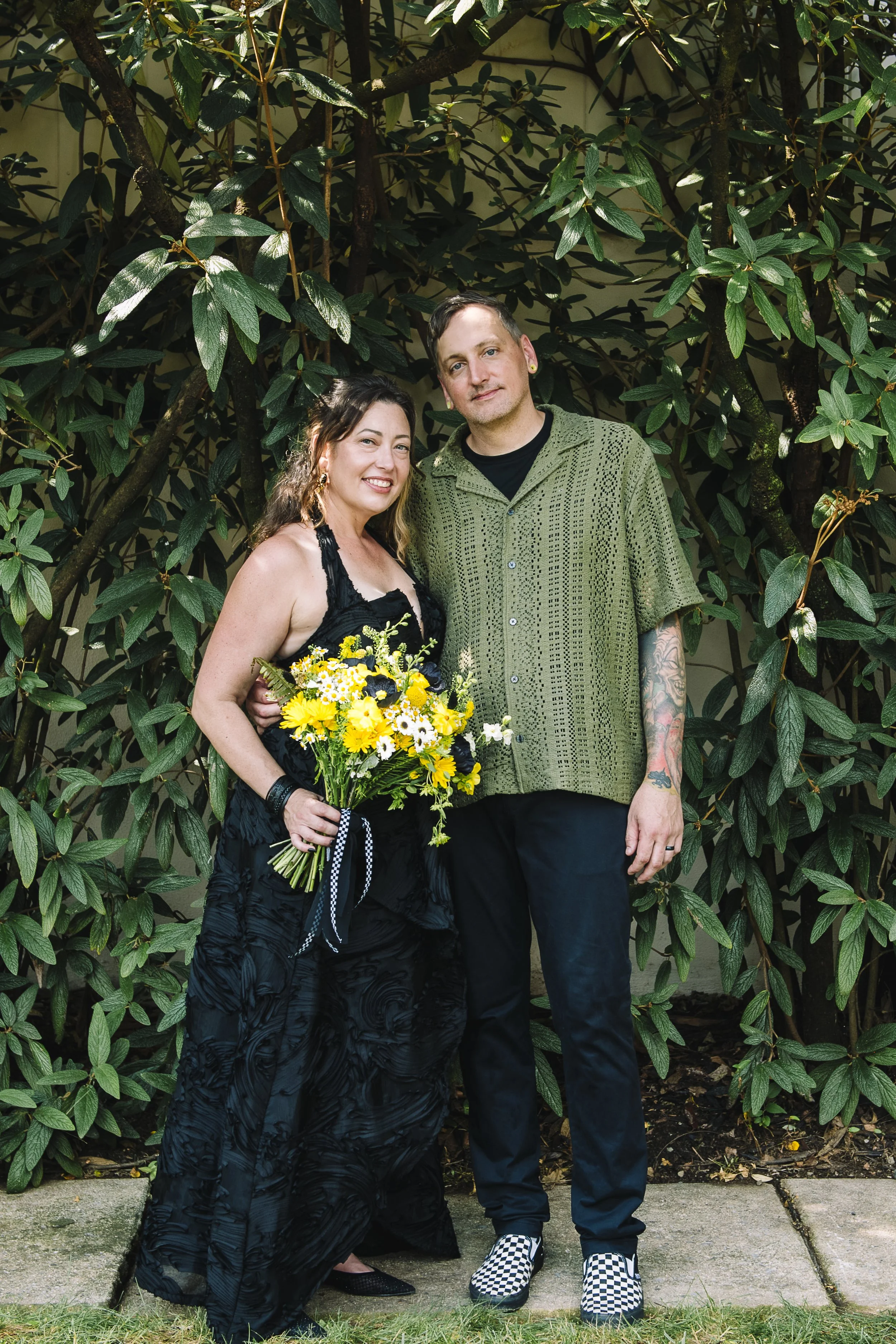 A woman and a man standing together outdoors in front of a large leafy shrub. The woman is holding a bouquet of yellow and white flowers, wearing a black dress, and smiling. The man is wearing a green patterned shirt, black pants, and checkered shoes