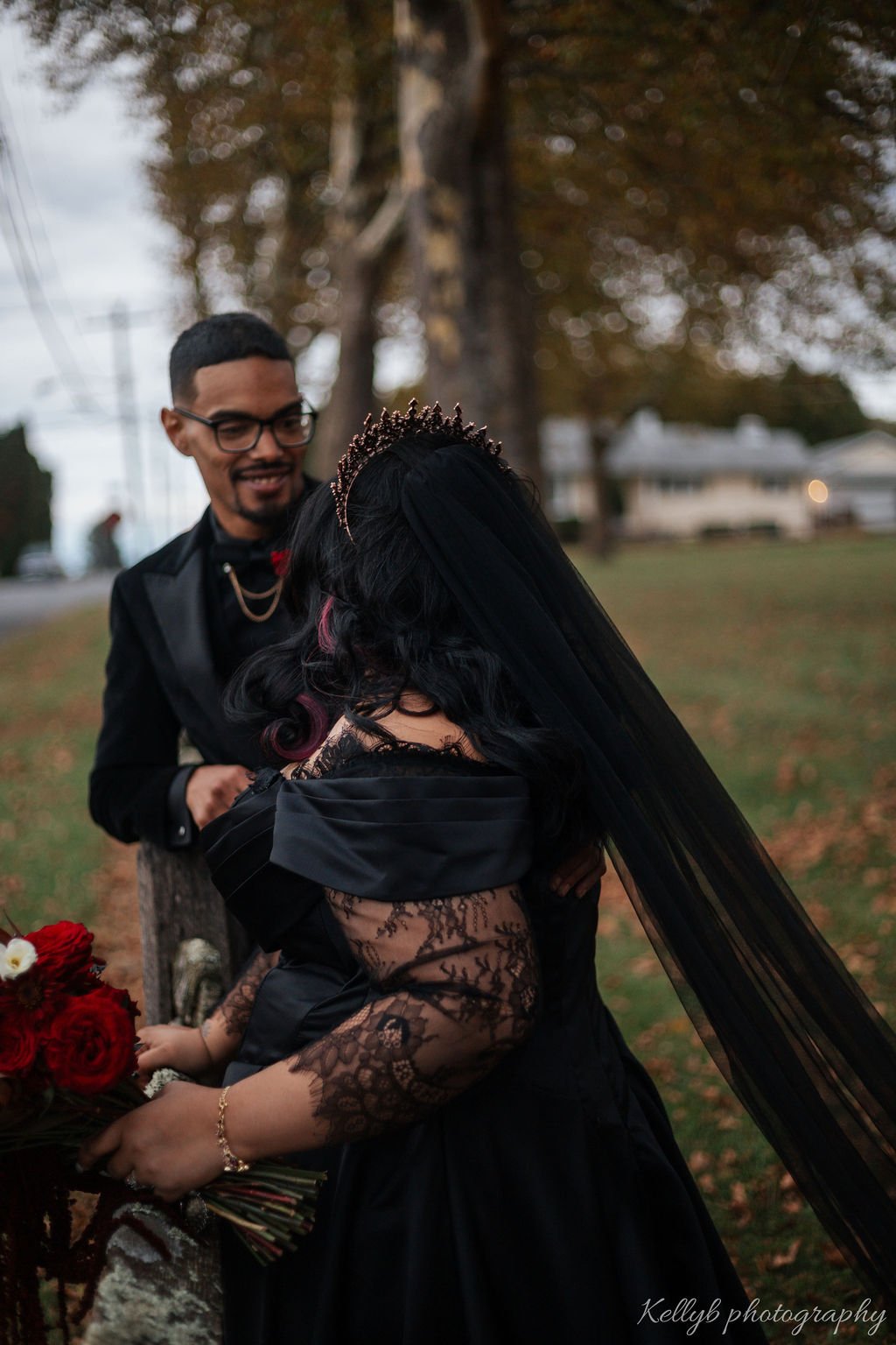 A couple dressed in formal attire, with the woman wearing a black lace dress and a tiara, and the man wearing a black suit, standing outdoors with trees and houses in the background.