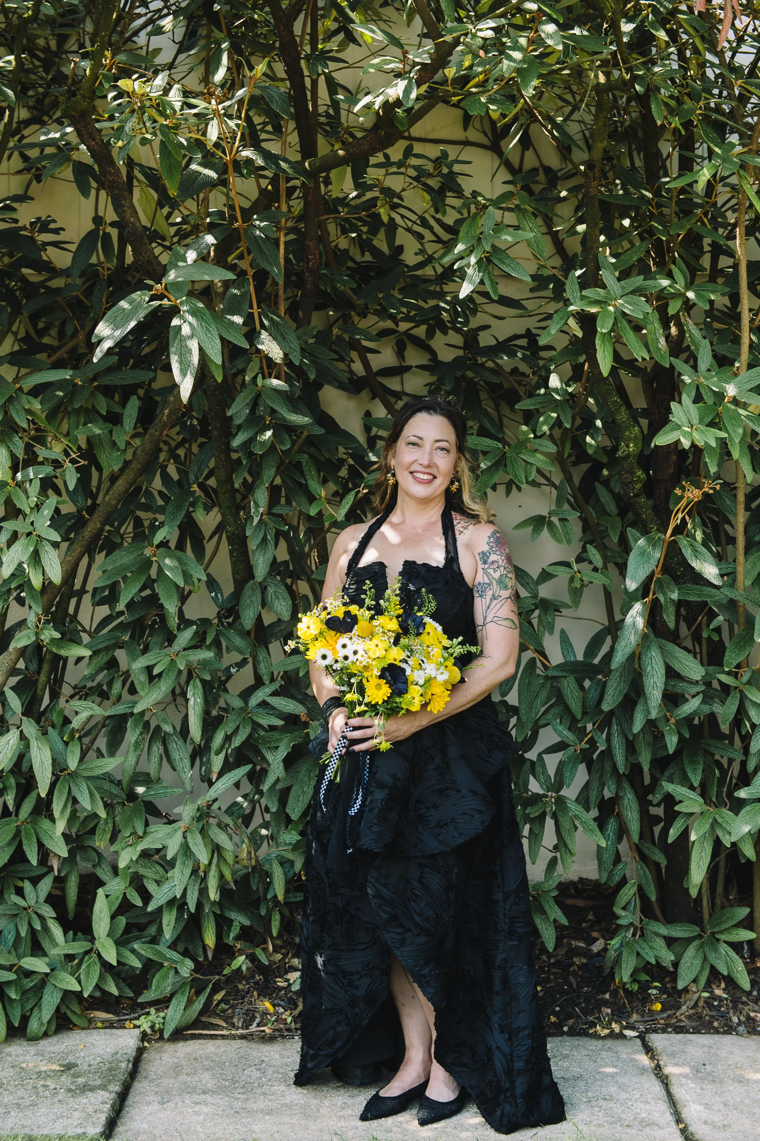 A woman in a black dress holding a yellow and white bouquet of flowers, standing outdoors in front of green foliage, smiling at the camera.