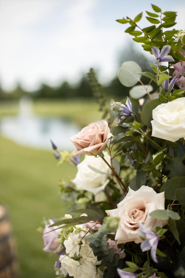 Close-up of a floral arrangement with white and blush roses, surrounded by green leaves and small purple flowers, outdoors with a grassy field and water in the background.
