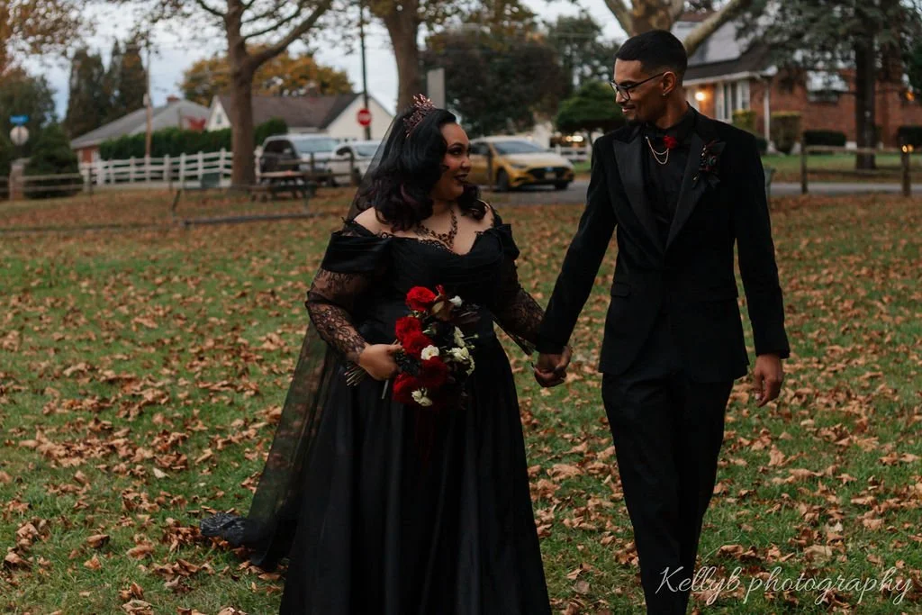 Couple walking hand in hand outdoors on a fall day, with the woman holding a bouquet of red and white flowers, dressed in a black gown with lace details, and the man in a black suit with a red bow tie, on a leaf-covered grass field.