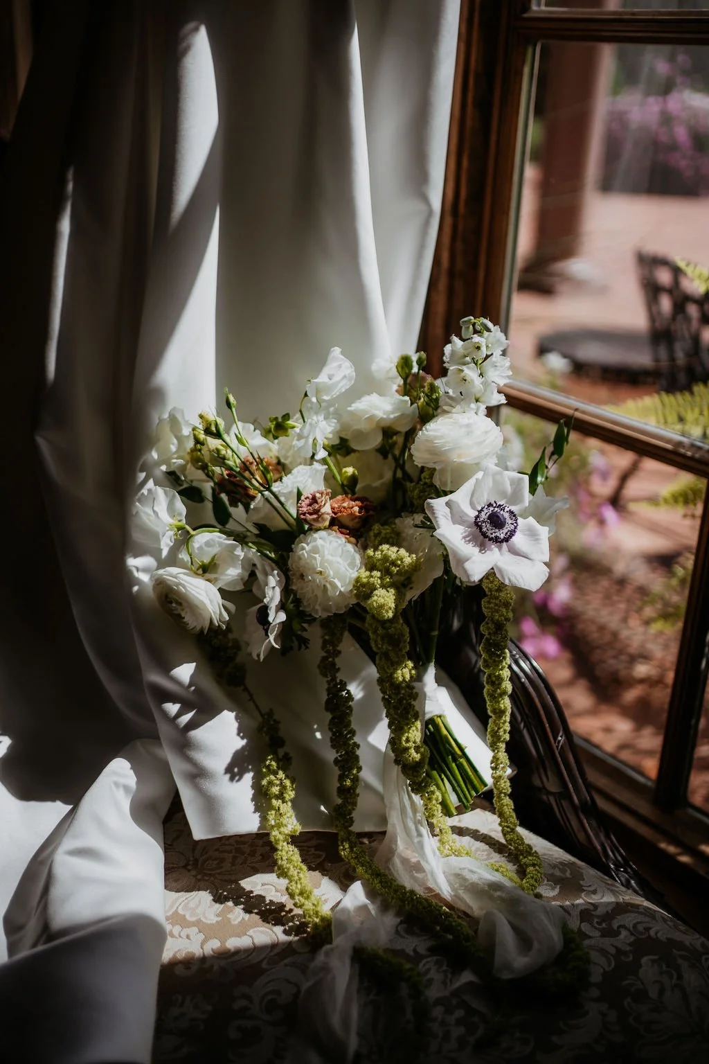 A bouquet of white and soft pink flowers resting on a table near a window with sunlight streaming in, with a white cloth draped around the flowers and a garden outside.