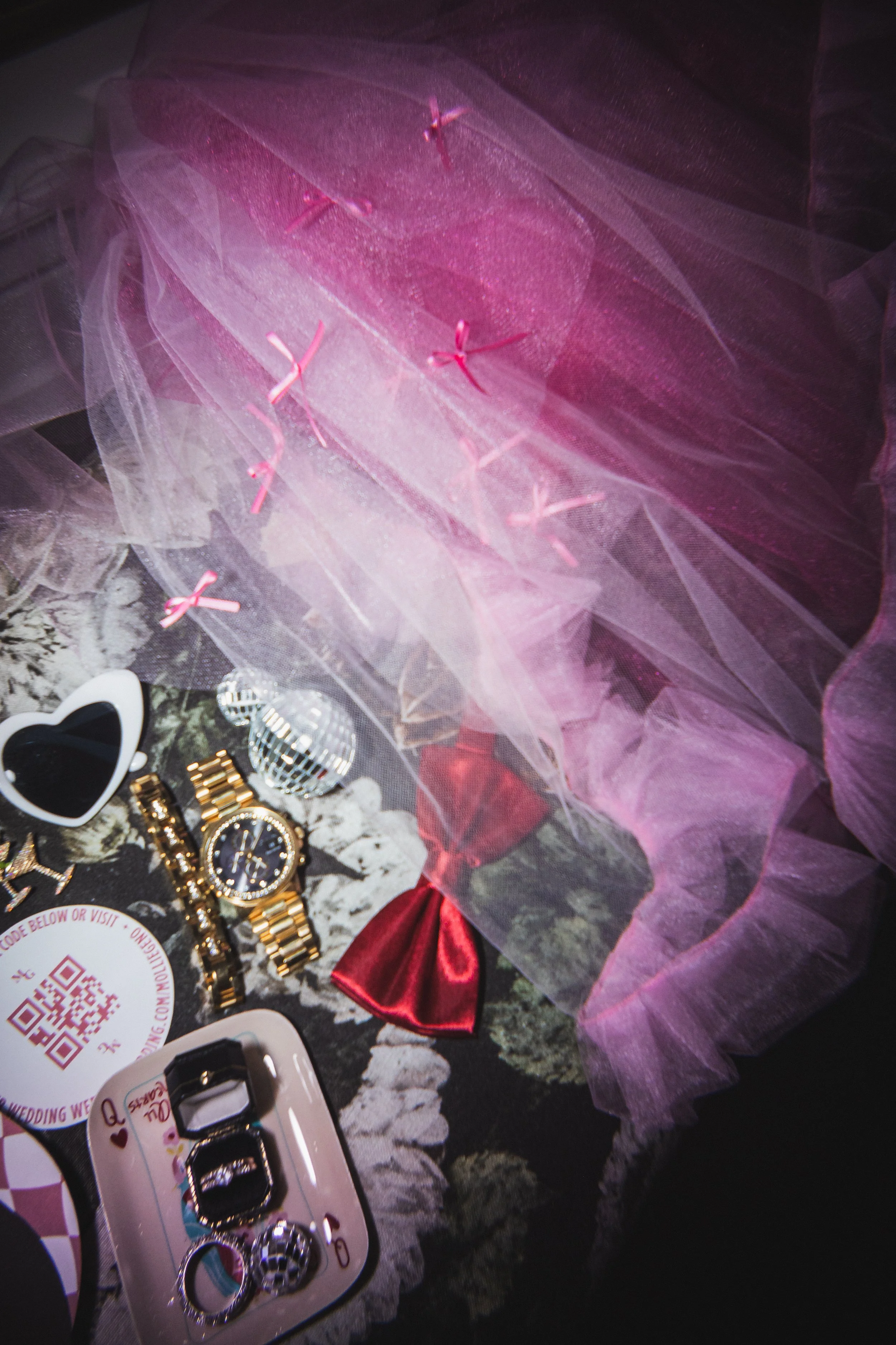 Assorted jewelry including a gold watch, rings, and earrings, a pink satin bow, a disco ball, and a pink tulle fabric with pink ribbons, all arranged on a floral patterned surface.