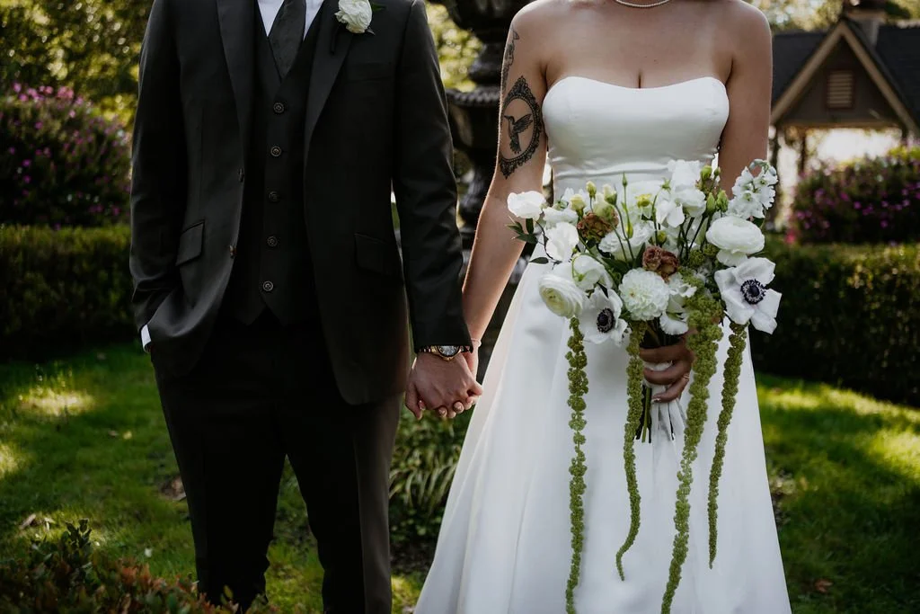 A bride and groom holding hands outdoors during a wedding, with bride in a strapless white dress and holding a bouquet of flowers, and groom in a black suit, in a garden setting.