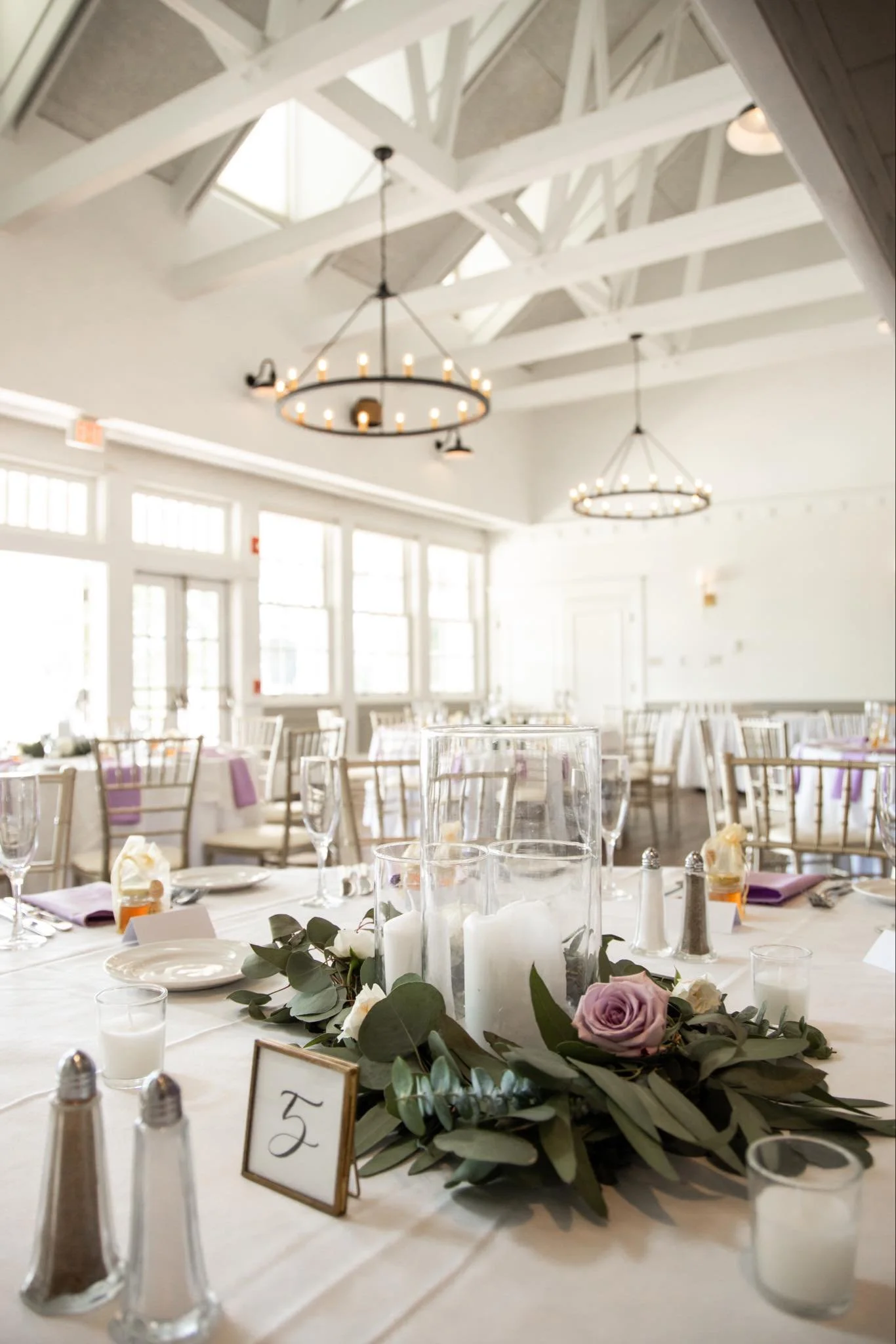 Elegant wedding reception table setup with purple roses, white candles, glassware, and purple napkins in a bright, airy room with high ceilings and chandeliers.