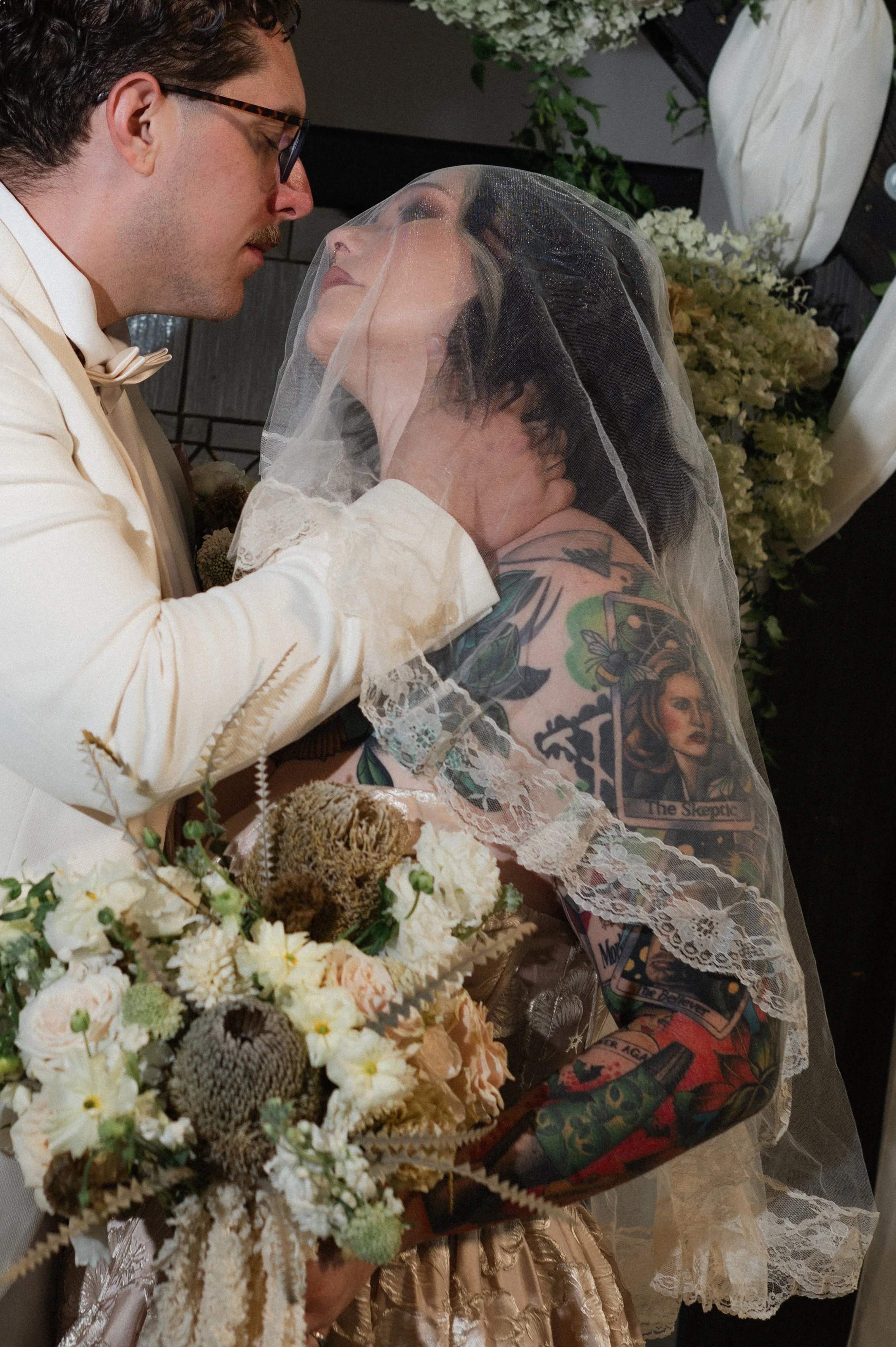 A groom and bride share an intimate moment during their wedding ceremony, with the groom gently holding the bride's face under a veil. The bride has visible tattoos on her arms and shoulders, and the groom wears glasses and a white suit with a bow ti