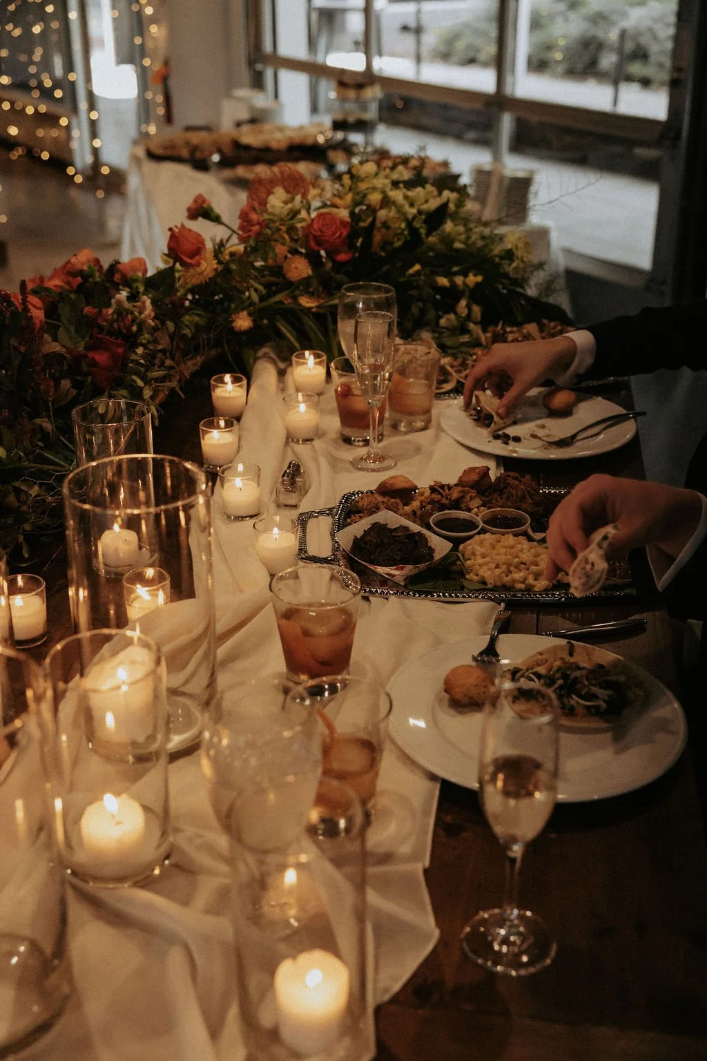 A formal dinner table decorated with candles and flowers, set with plates of food, drinks, and silverware, in a dimly lit room with large windows.