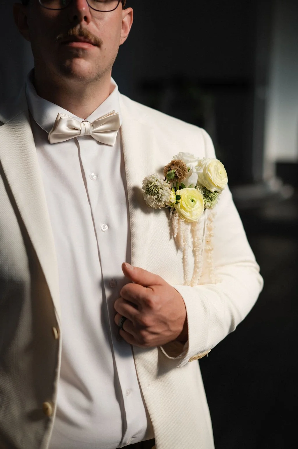 A man dressed in a white tuxedo with a bow tie, holding the lapel of his jacket with one hand, displaying a boutonniere of white and cream flowers on his chest.