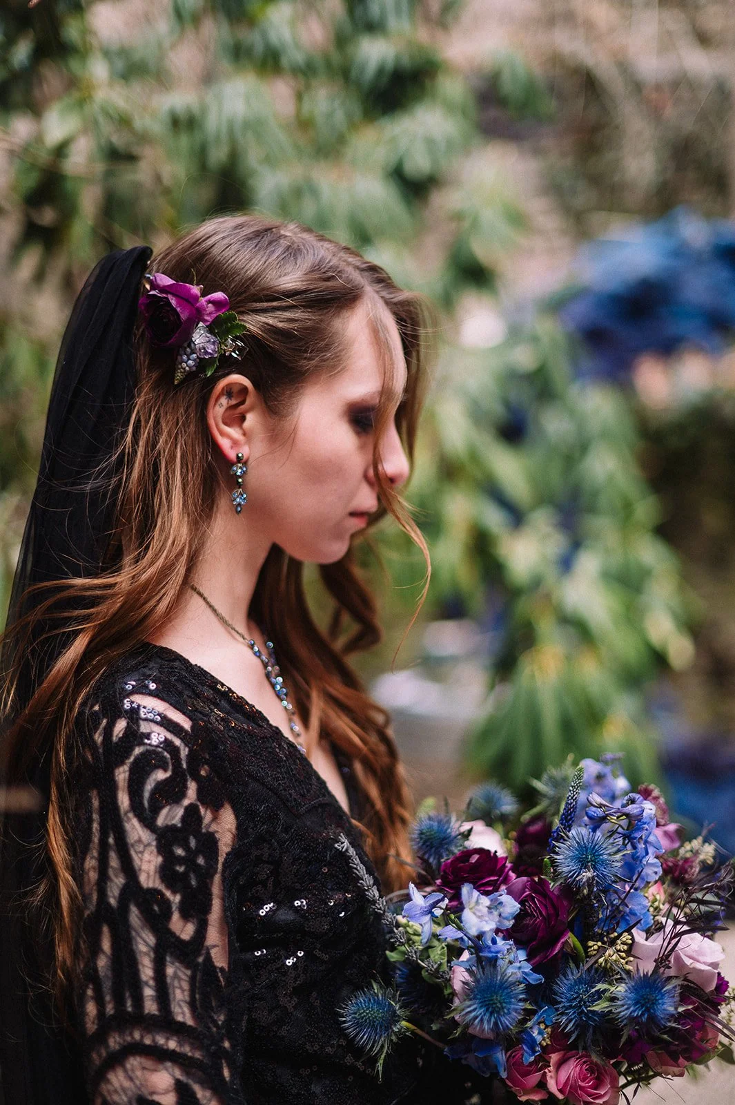 A young woman in black lace dress holding a bouquet of purple, pink, and blue flowers, with purple flowers in her hair, standing next to greenery.