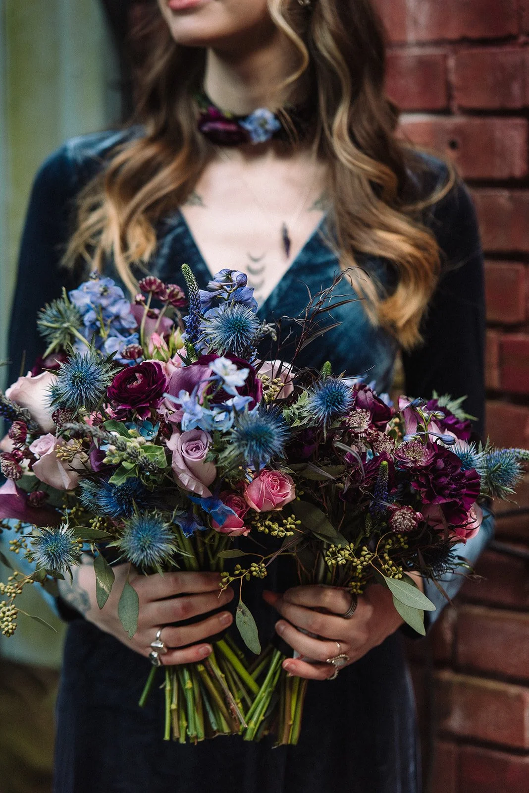 A woman holding a large bouquet of purple, pink, and blue flowers in front of a brick wall.