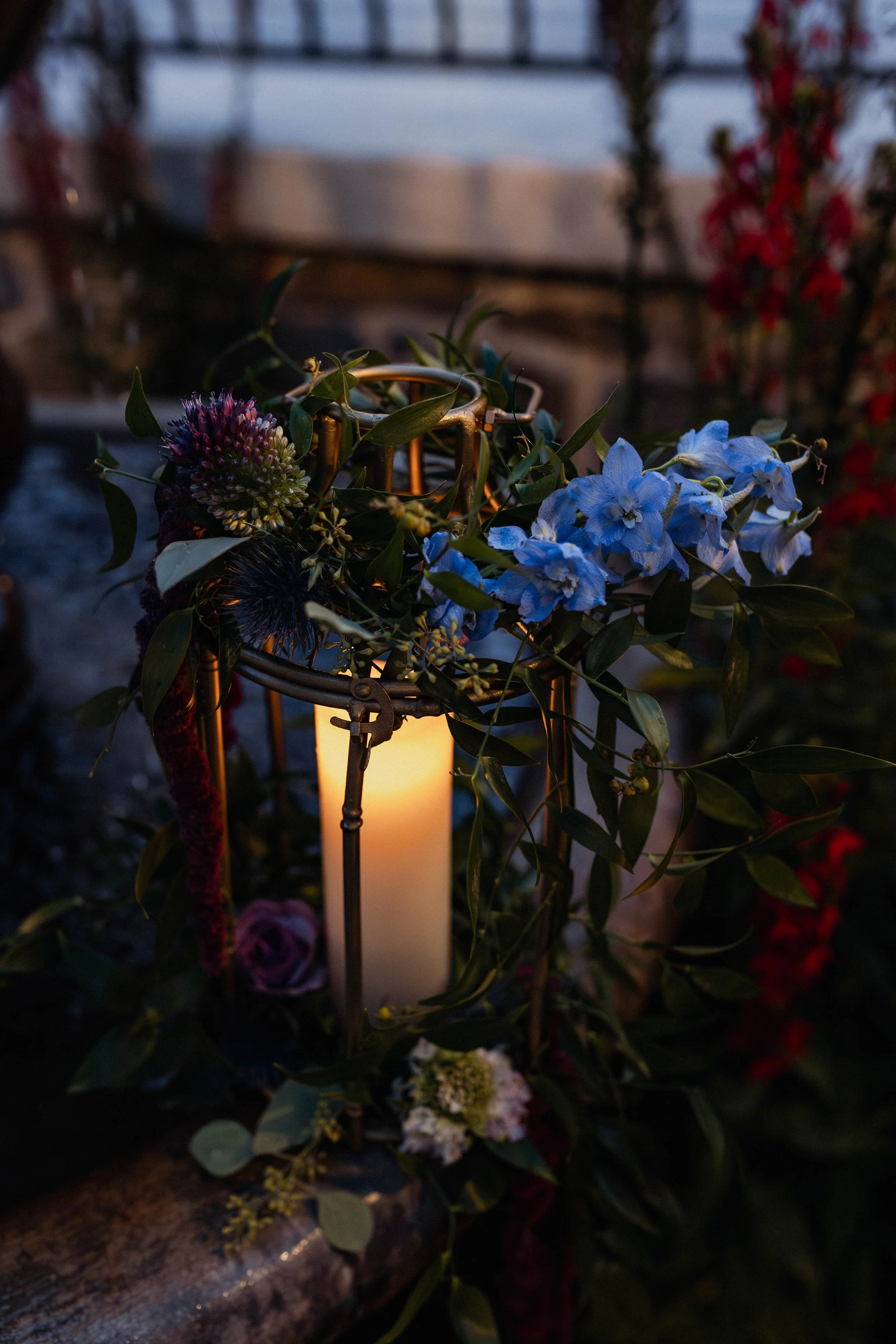 A candle inside a lantern surrounded by flowers and greenery, set outdoors during the evening or night.
