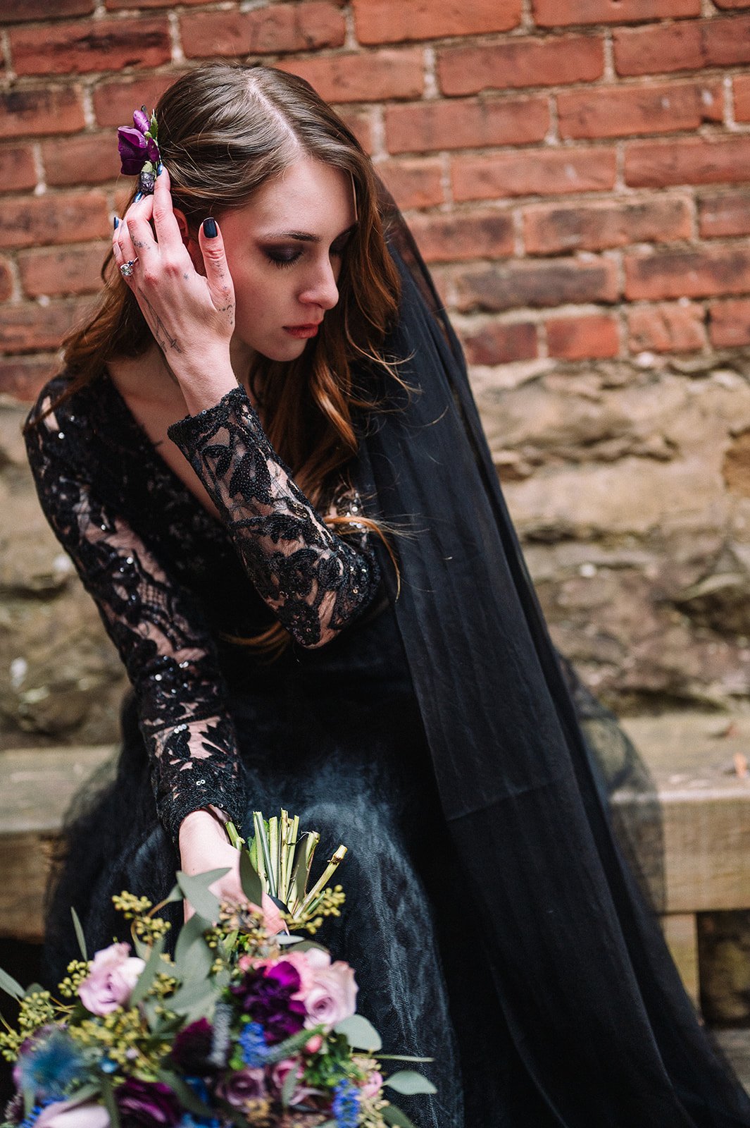 A woman with long wavy brown hair, wearing a black lace dress, sitting next to a brick wall, holding a bouquet of colorful flowers, with her head slightly bowed and touching her ear.