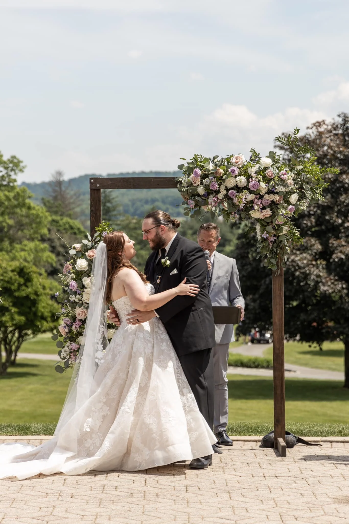 A bride and groom share a kiss during an outdoor wedding ceremony, with an officiant standing behind them, under a floral arch with pink, white, and purple flowers. Green trees and a partly cloudy sky are in the background.